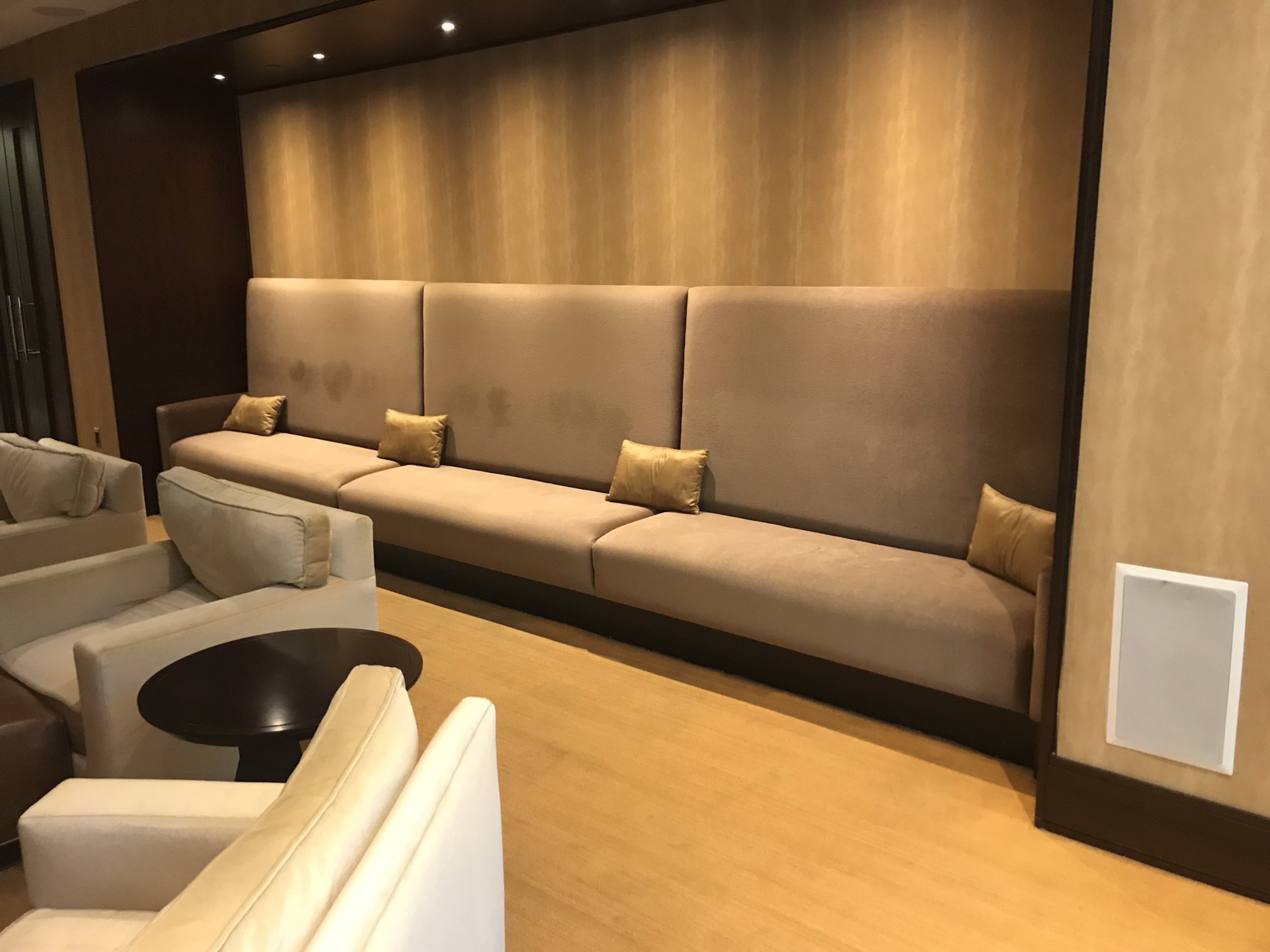 A modern seating area featuring a long beige upholstered bench with several small pillows against a textured wall, accompanied by soft, light-colored armchairs and a dark round table.