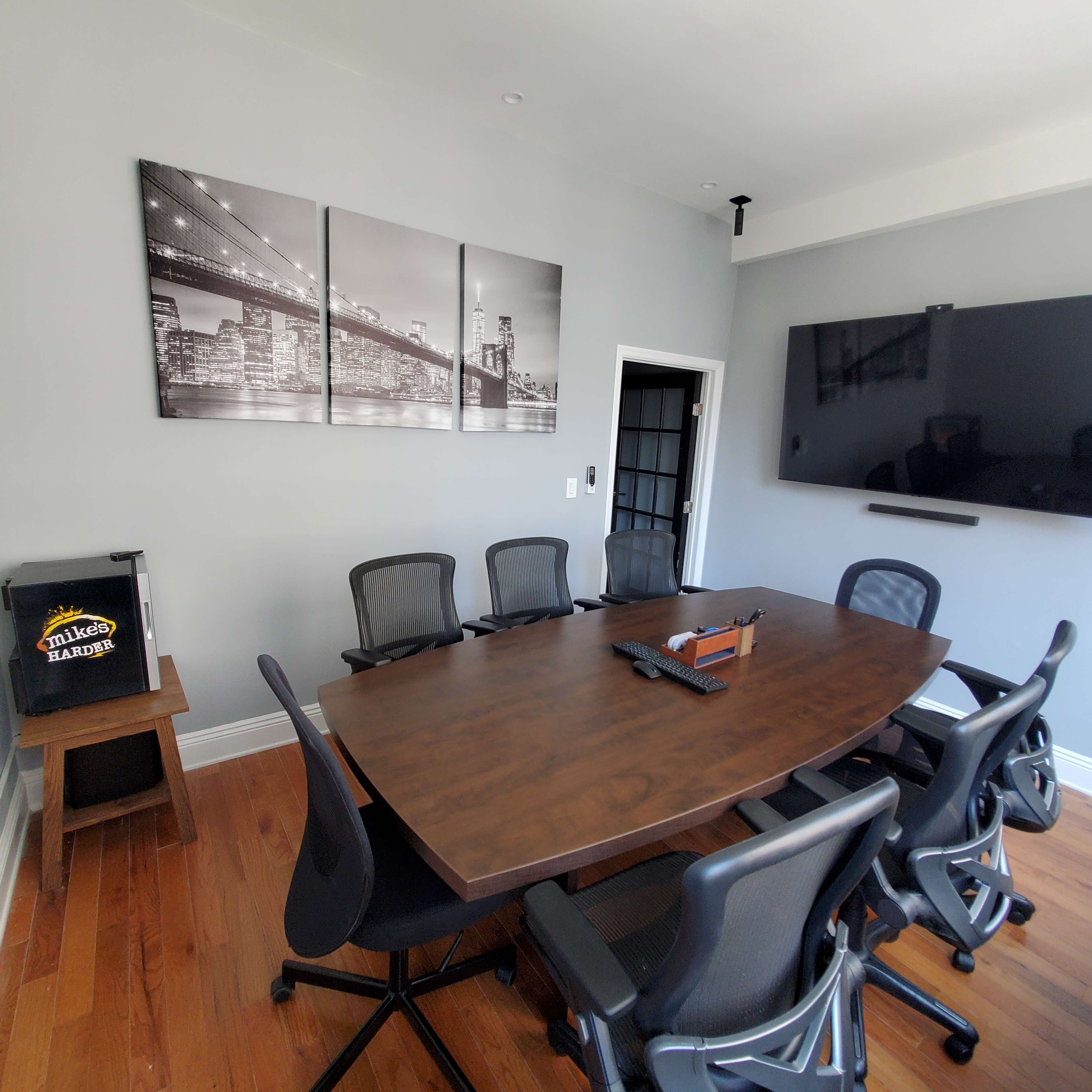 A conference room with a large wooden table surrounded by black chairs, featuring a wall-mounted TV and a set of black-and-white photographs of a city skyline.