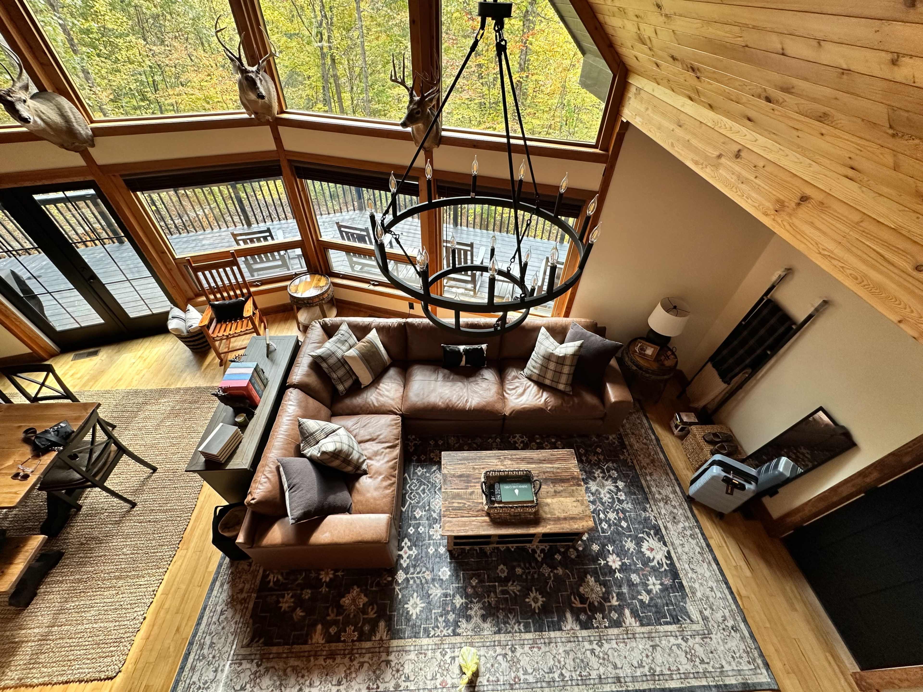 The image shows a spacious, modern living room with a large brown leather sectional sofa, a wooden coffee table, and high ceilings adorned with a circular chandelier.