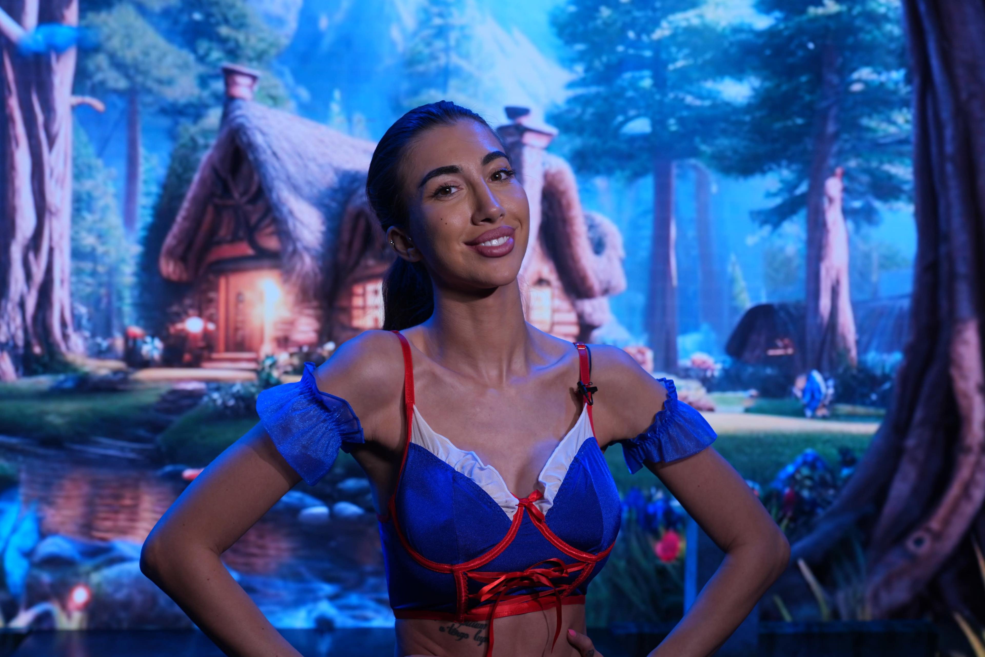 A woman poses in front of a colorful, animated forest backdrop featuring a quaint cottage.