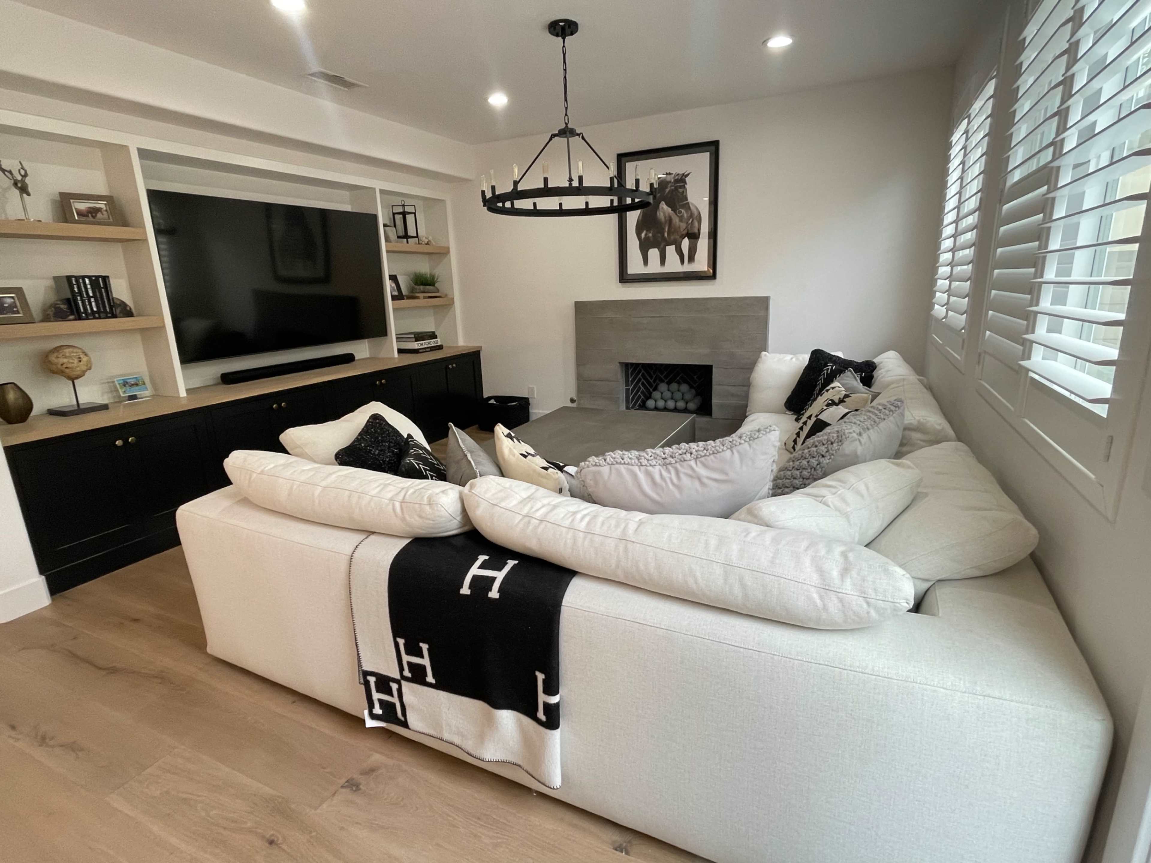 A large, light-colored sectional couch with black and white decorative pillows is positioned in a modern living room with a television, a stone fireplace, and built-in shelving against the wall.