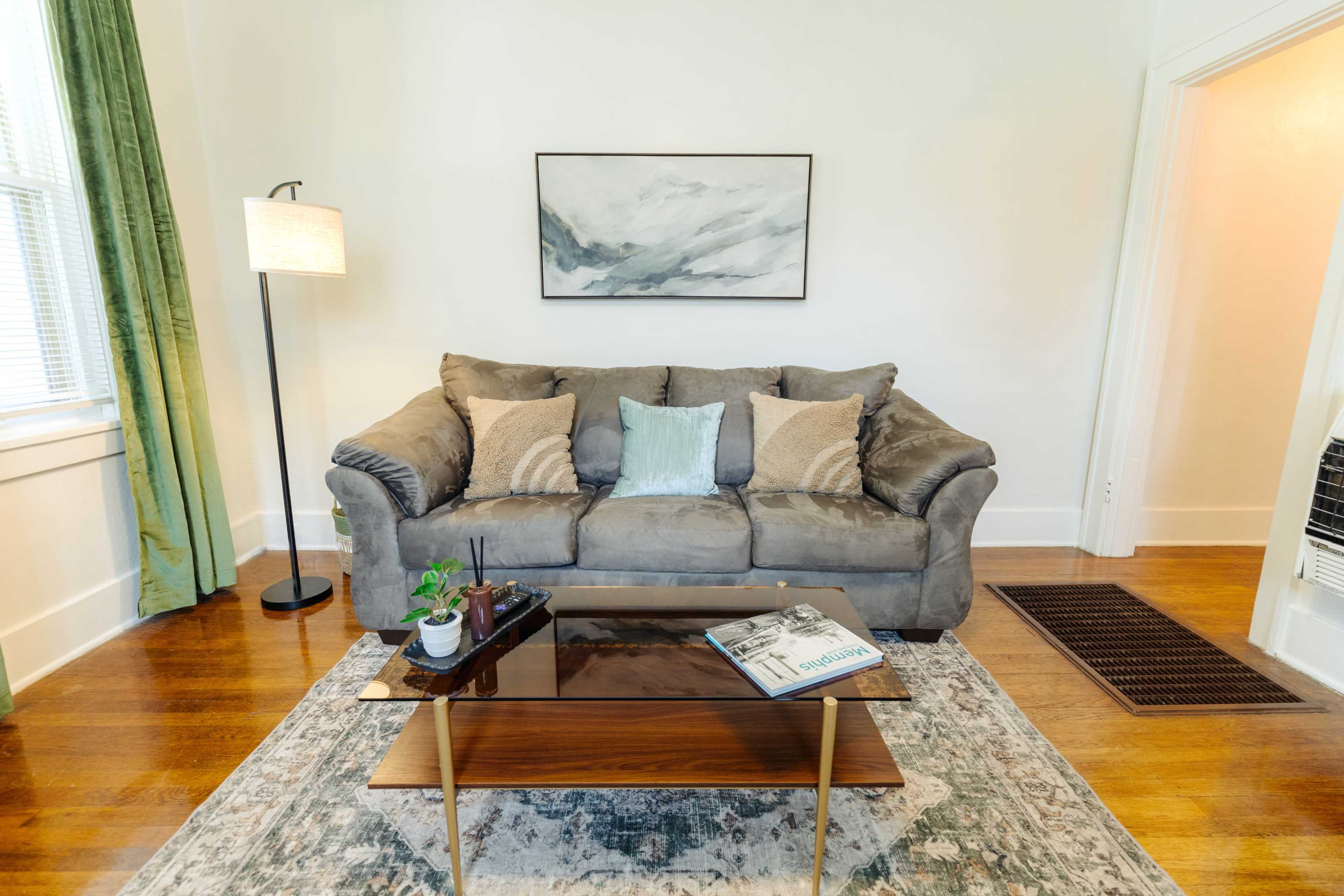 A modern living room features a gray sofa with decorative pillows, a wooden coffee table with a magazine, and a standing lamp beside large windows with green curtains.