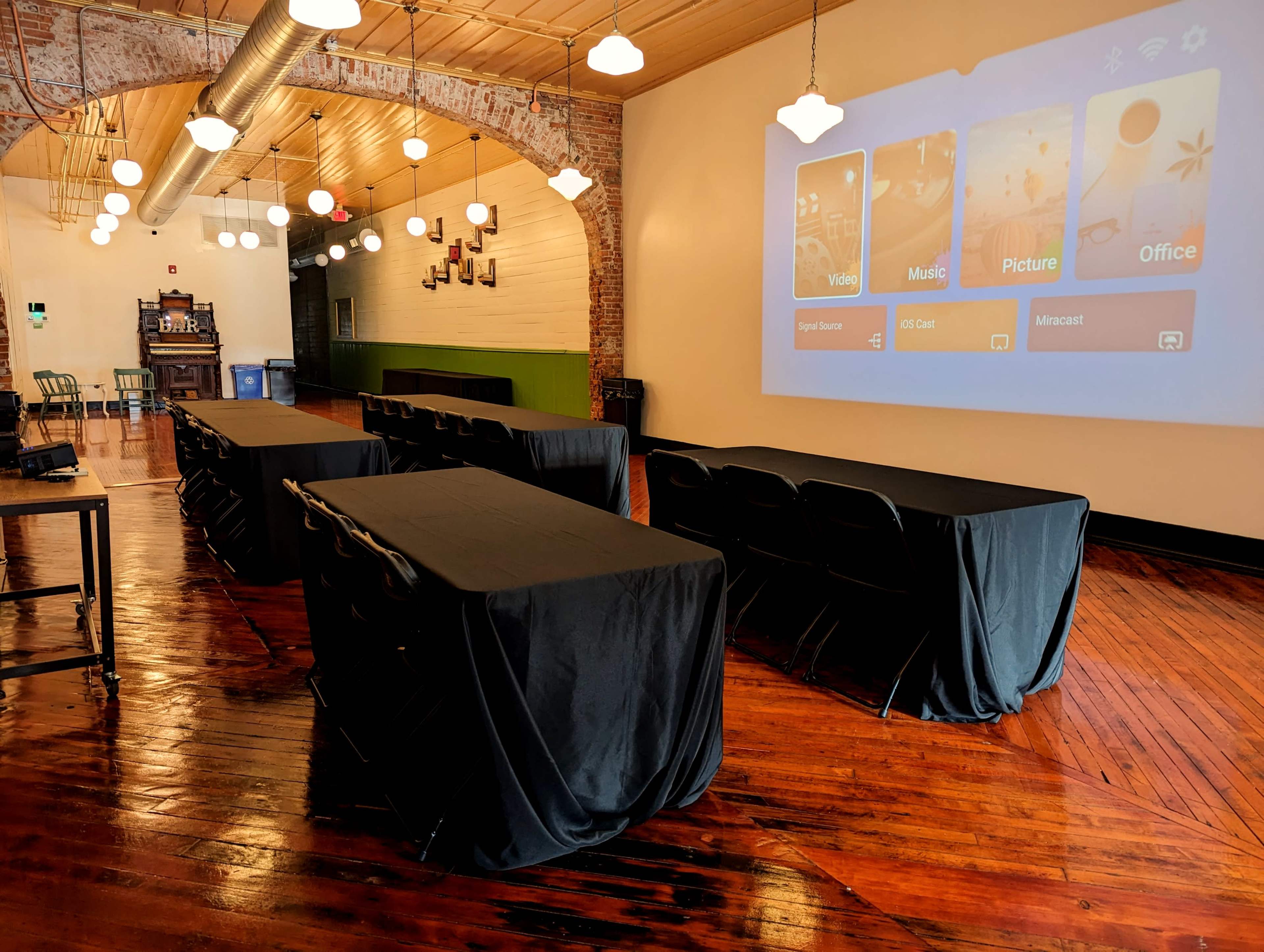 A spacious room with several rows of covered tables, a large screen displaying multimedia options, and pendant lighting hanging from the ceiling.