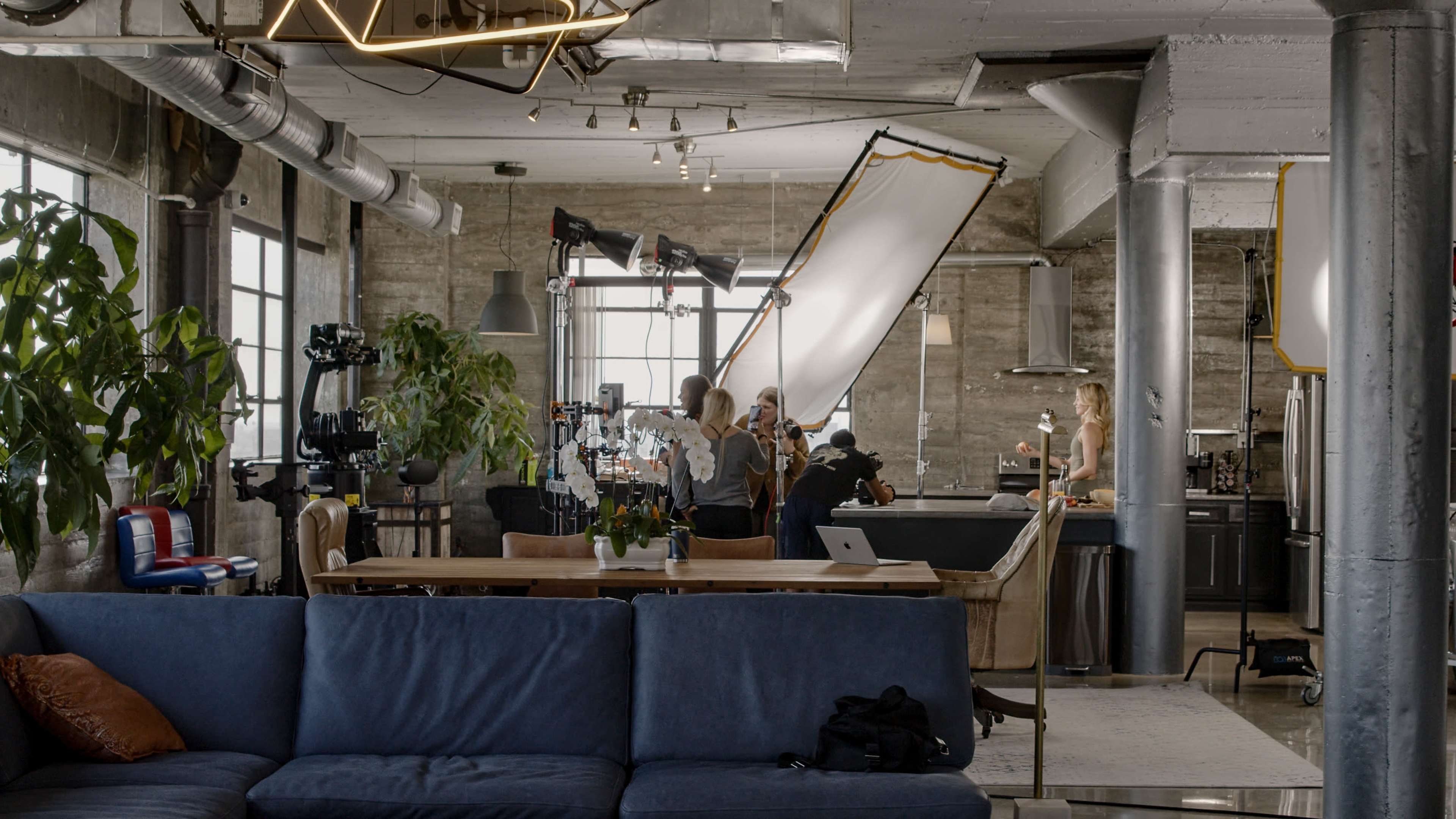 A modern studio space features a dining area, plants, and production equipment with a film crew working on a shot.