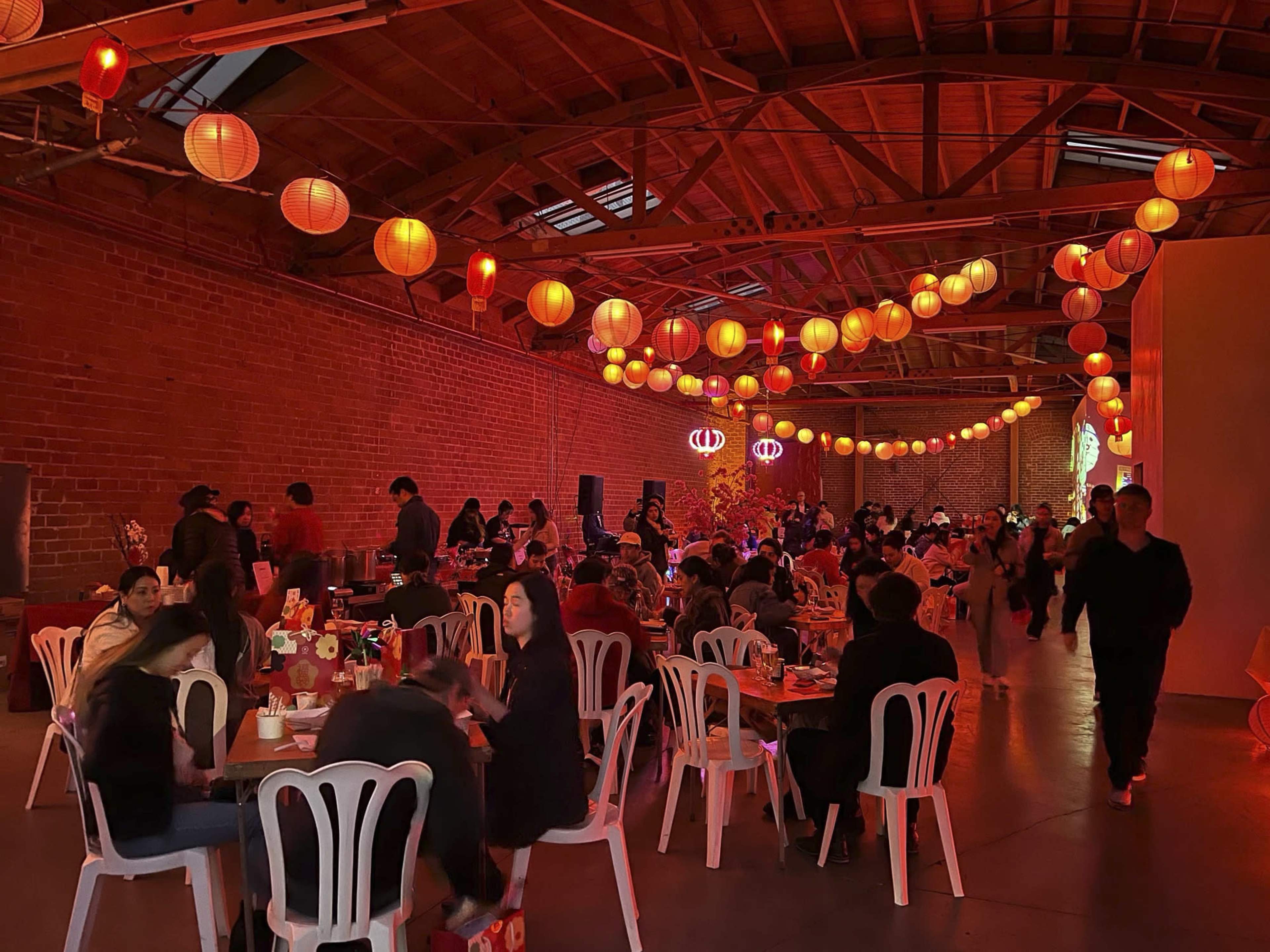 A festive gathering takes place in a large, open-space venue decorated with orange lanterns hanging from the ceiling, where tables are filled with guests enjoying food and conversation.