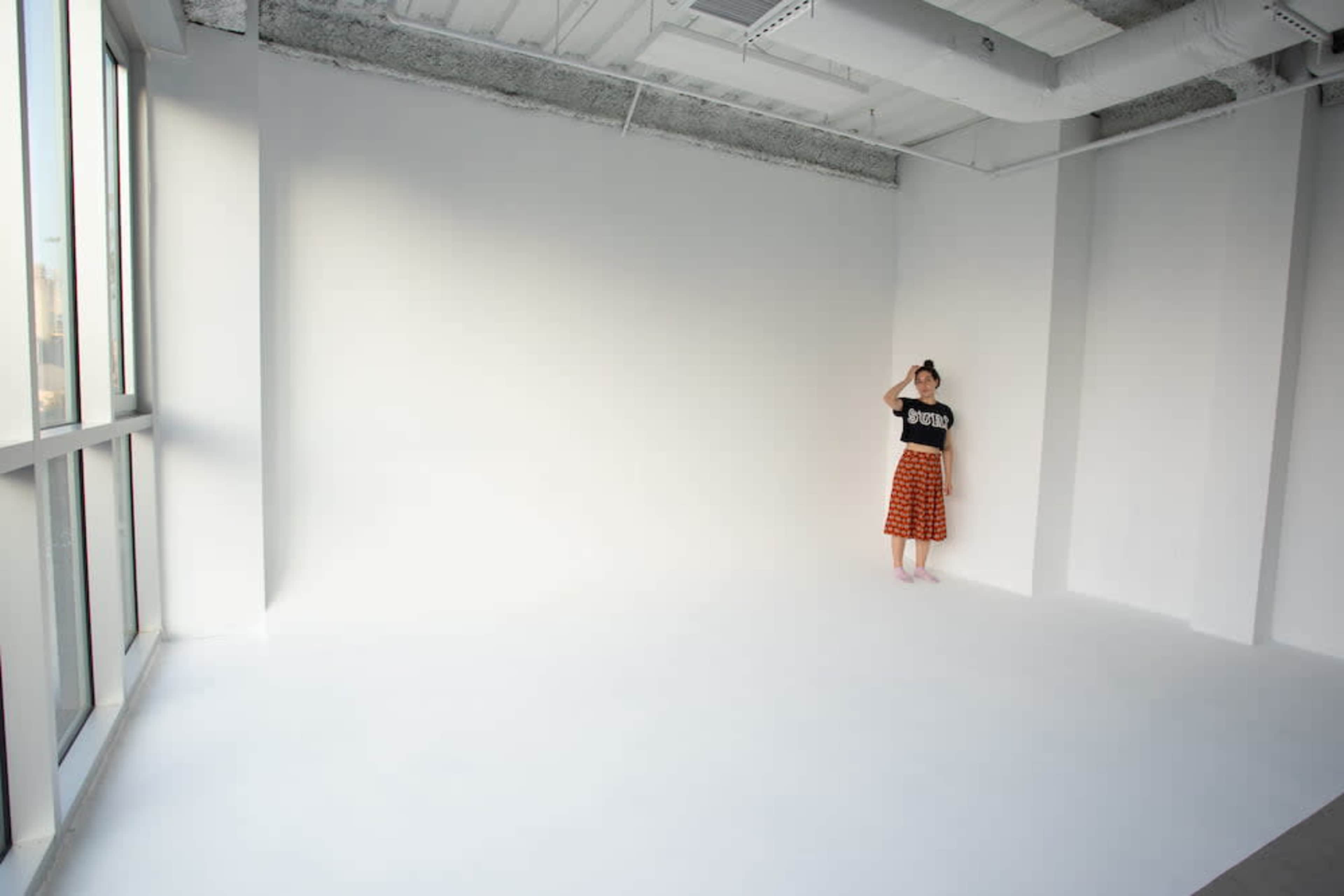A person stands in a spacious, minimally decorated room with white walls and a large window.