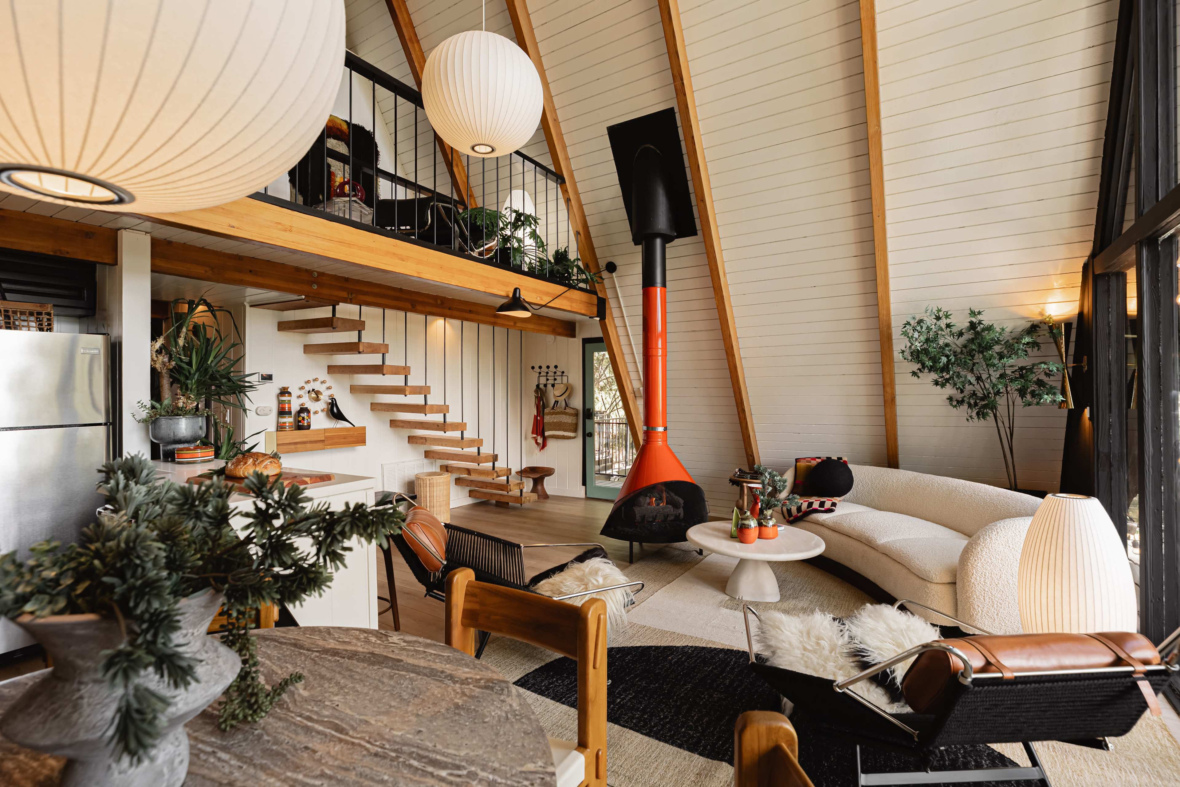 The image shows a modern, open-concept living room with a high ceiling, a staircase, and a prominent red fireplace, surrounded by various plants and stylish furniture.