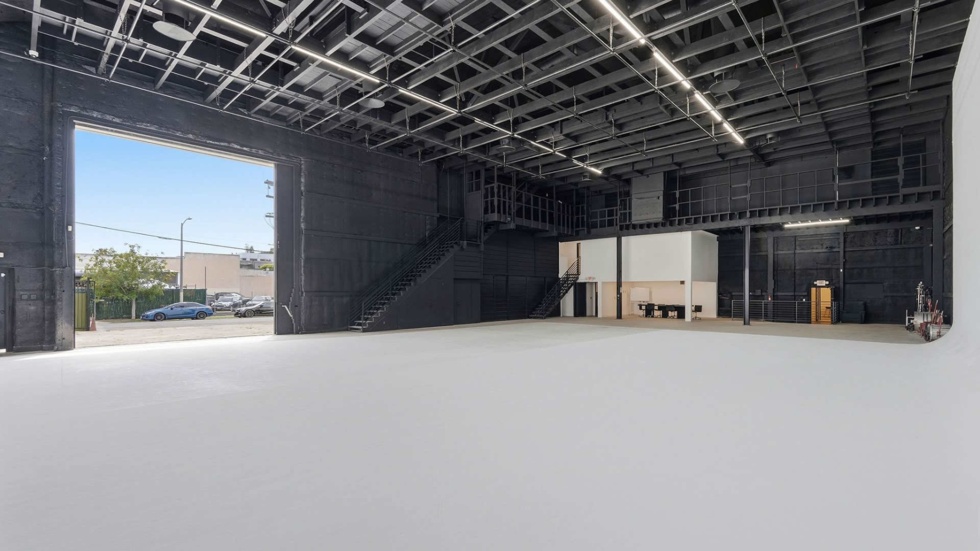 The image depicts an expansive, empty studio space with high ceilings, a large open door leading outside, and a staircase on the left side.