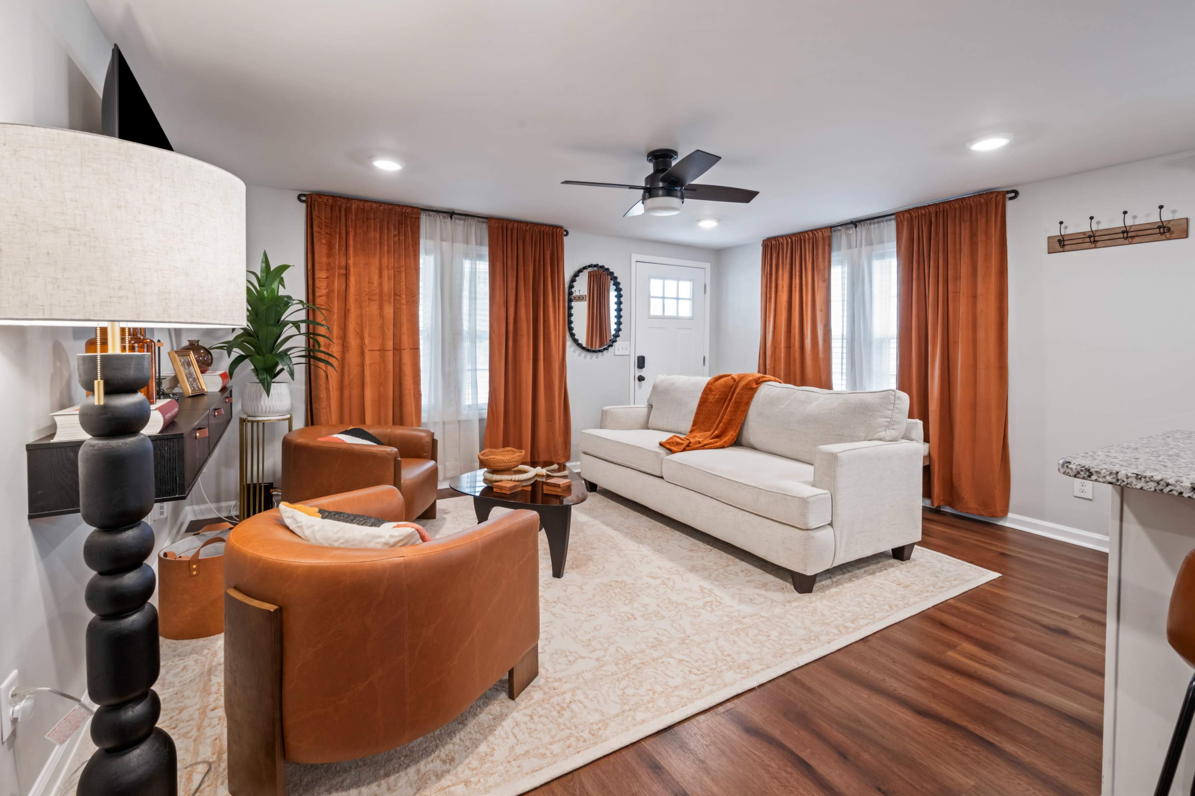 The image shows a cozy living room featuring a light-colored sofa, two brown leather chairs, orange curtains, and a patterned area rug, with a door leading outside and a ceiling fan above.