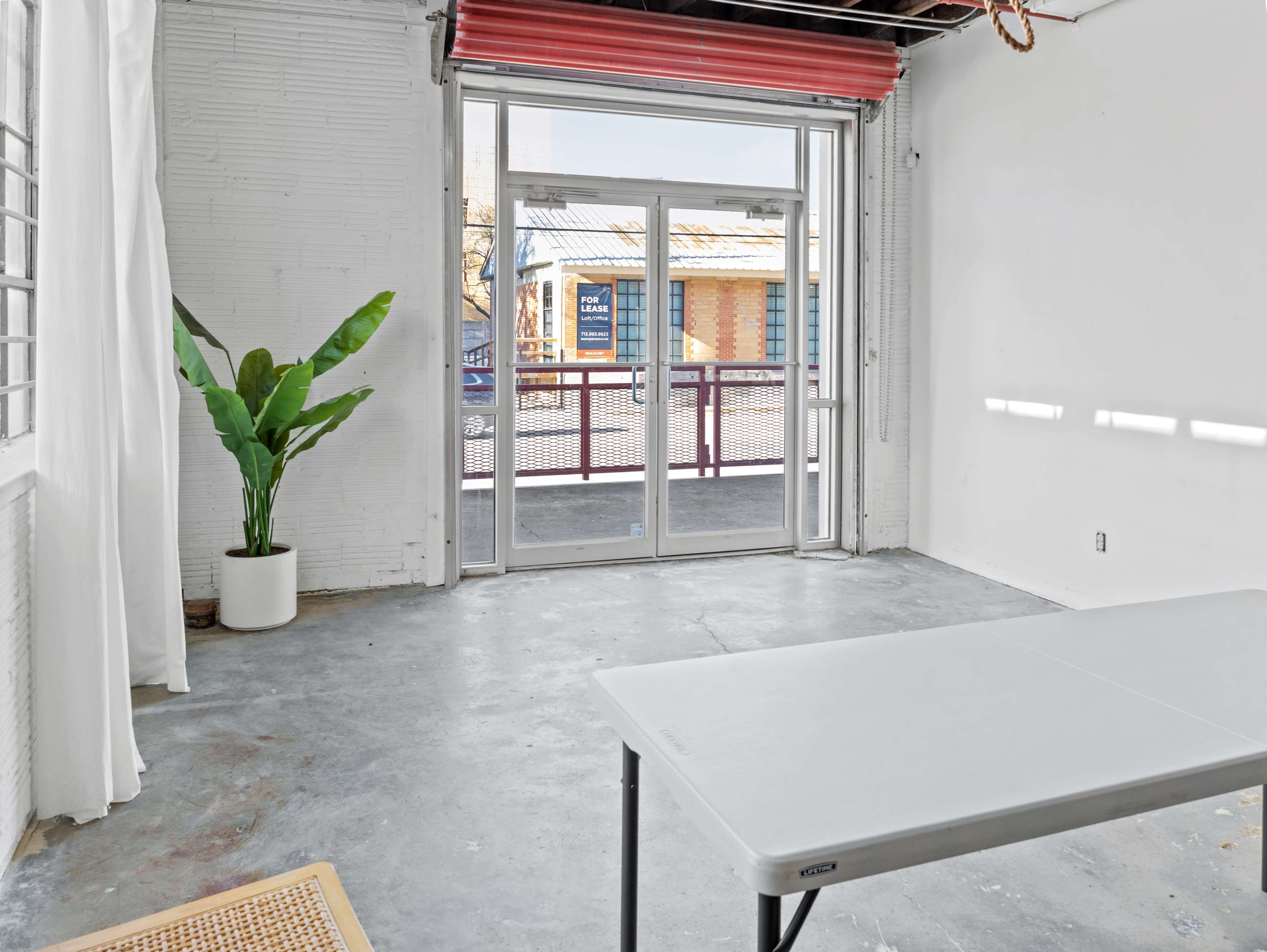 A minimalist interior room with a folding table, a large plant in a white pot, and a glass door leading to an outdoor area.