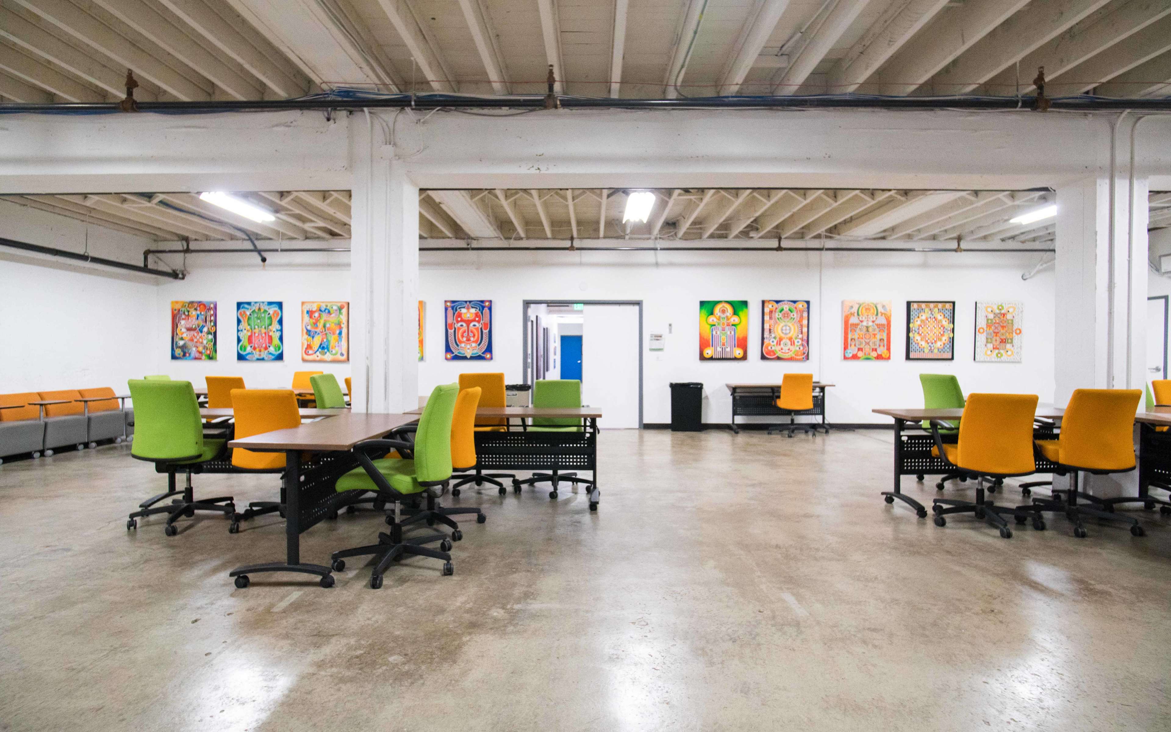 A spacious office area with several tables and chairs in green and orange, surrounded by colorful wall art.