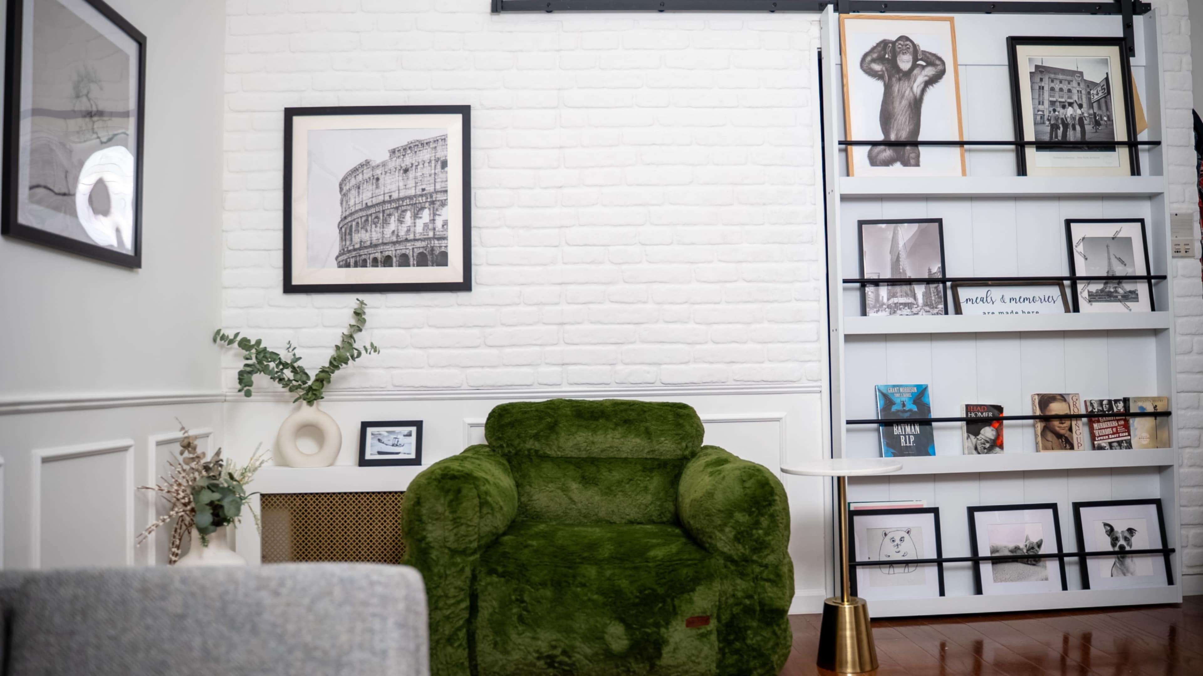 The image shows a cozy living room featuring a green armchair, a bookshelf with various artwork and books, and a framed photograph hanging on the wall.