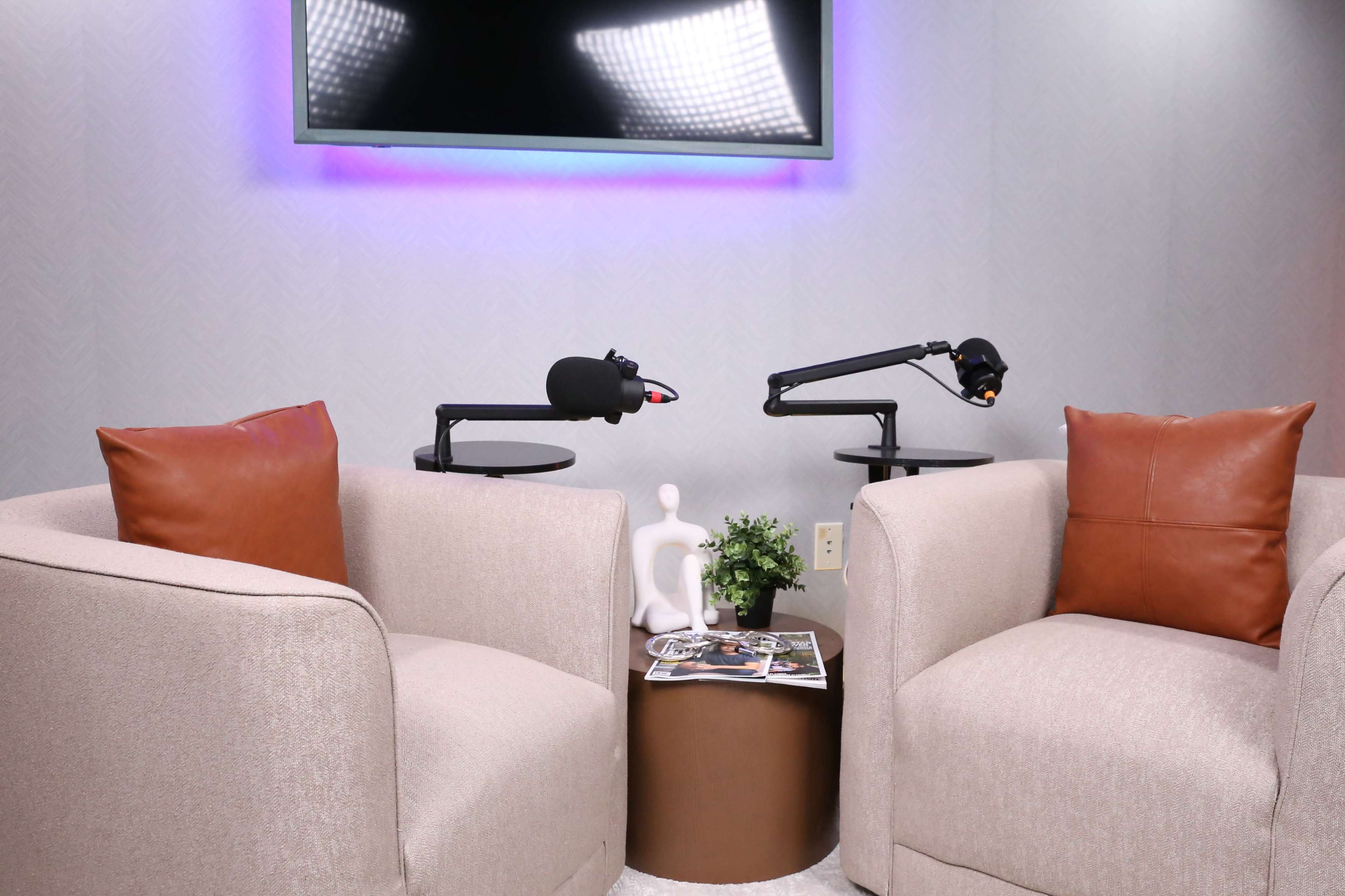 The image shows two beige armchairs with brown leather accents positioned around a small table, featuring a plant and magazines, in front of a wall-mounted screen.