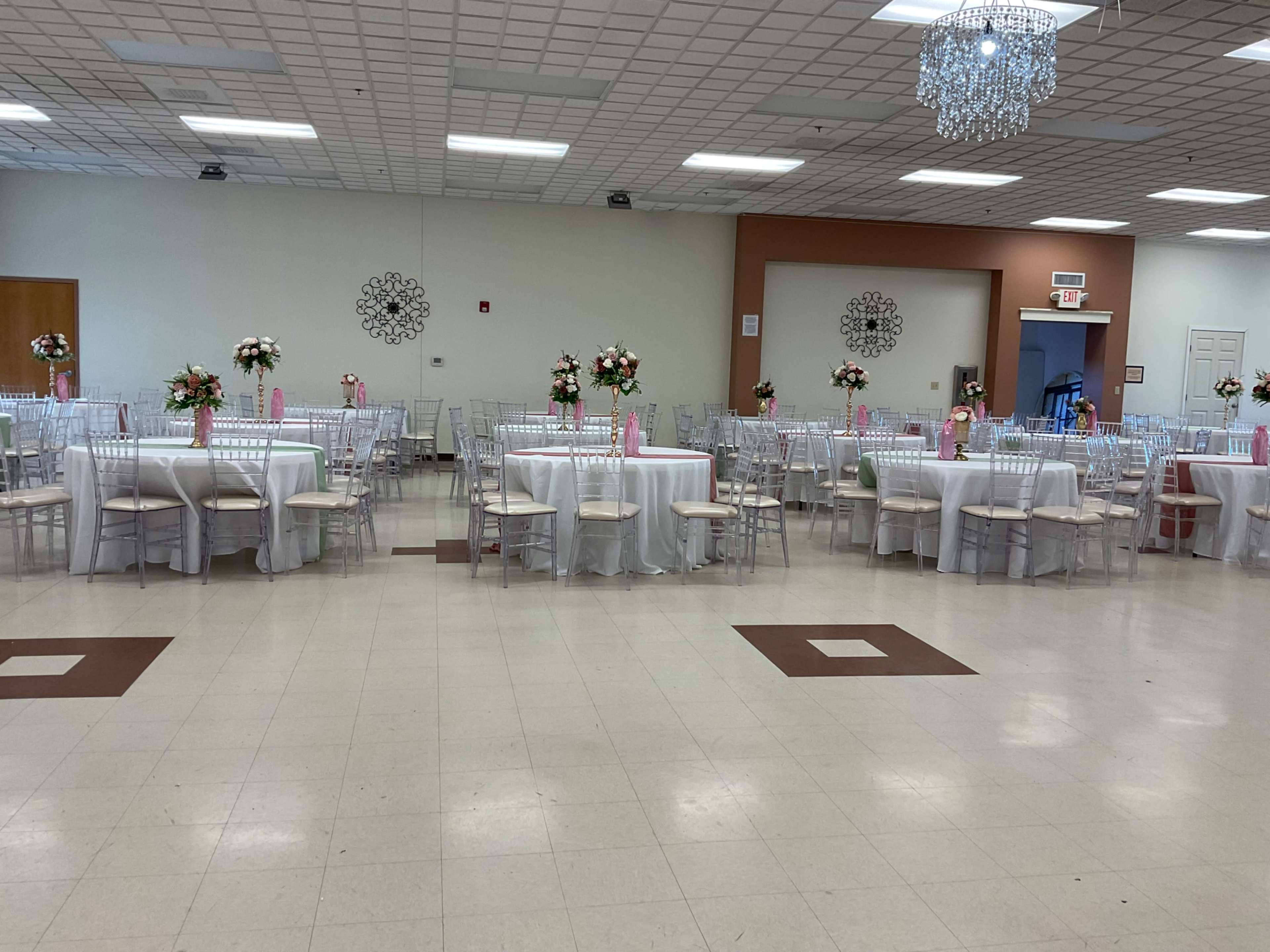 The image shows a spacious banquet hall set up for an event, featuring round tables with white tablecloths and floral centerpieces, arranged with clear chiavari chairs.