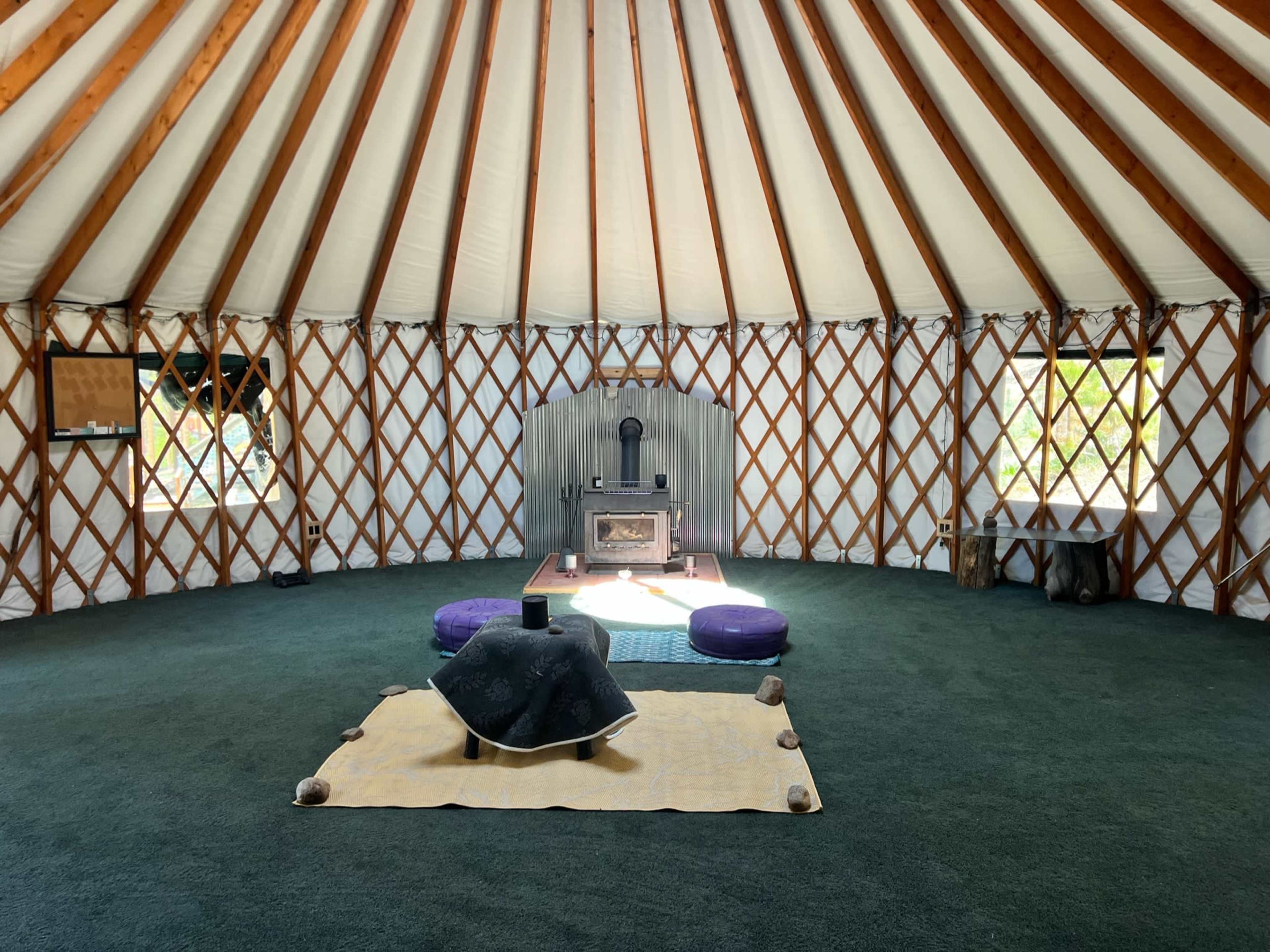The interior of a yurt features a circular layout with a central heating stove, circular seating arrangements, and a wooden framework.