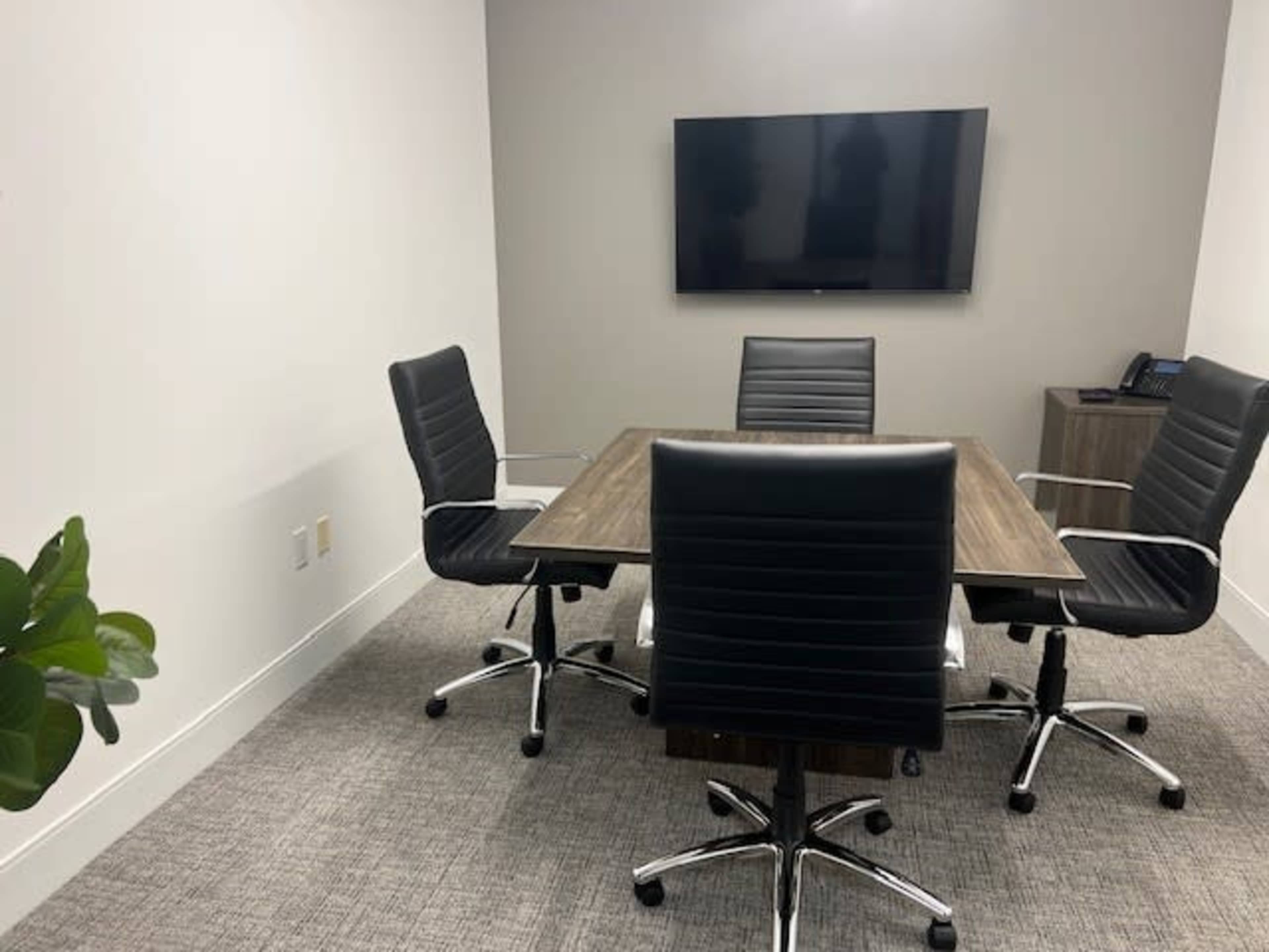 A conference room features a wooden table surrounded by four black leather chairs and a wall-mounted television.