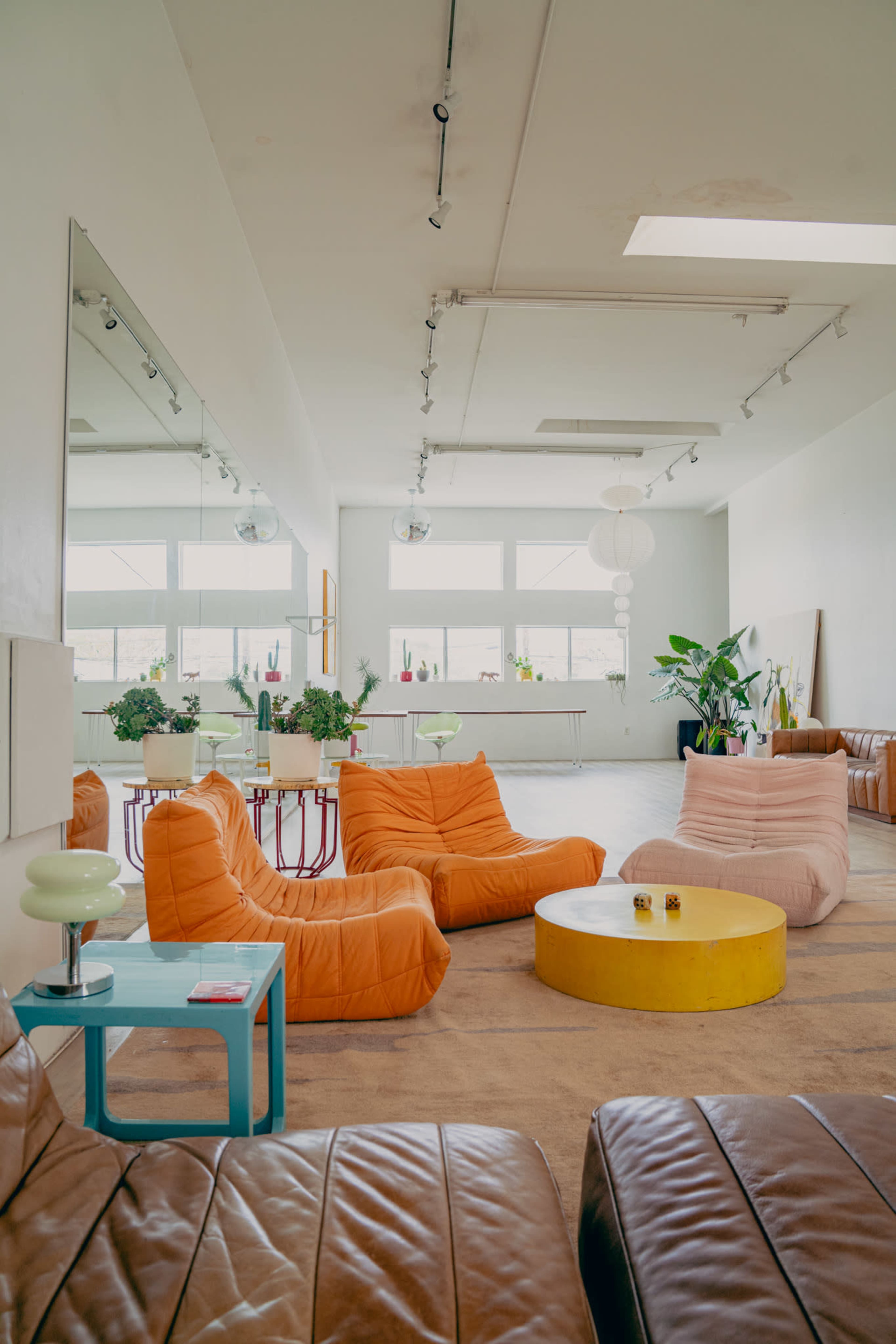 The image shows a bright, spacious room with eclectic furniture, including orange lounge chairs, a round yellow table, and various indoor plants.