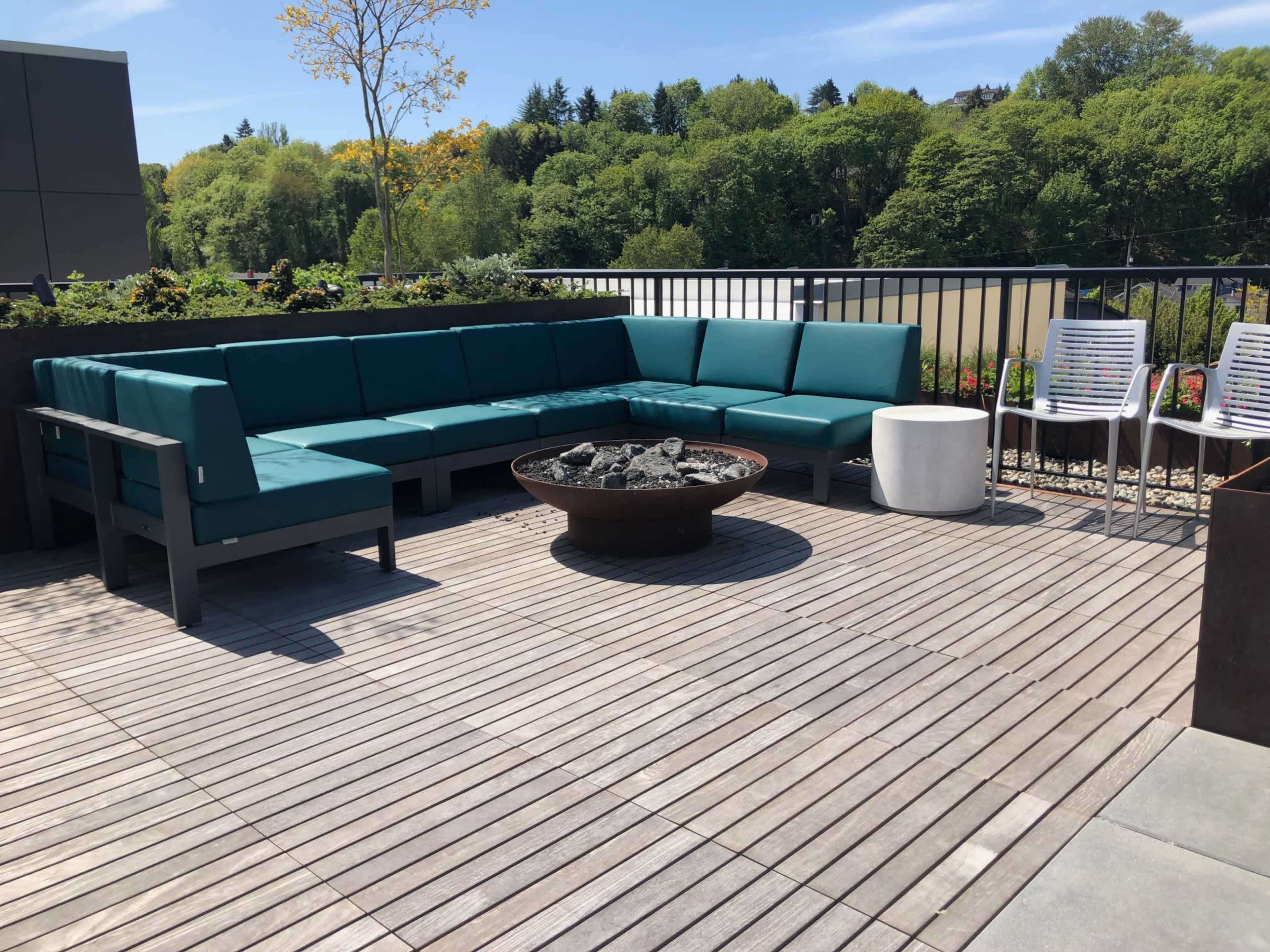 A modern outdoor seating area features a turquoise sectional sofa surrounding a round fire pit on a wooden deck, with a white table and additional chairs nearby.