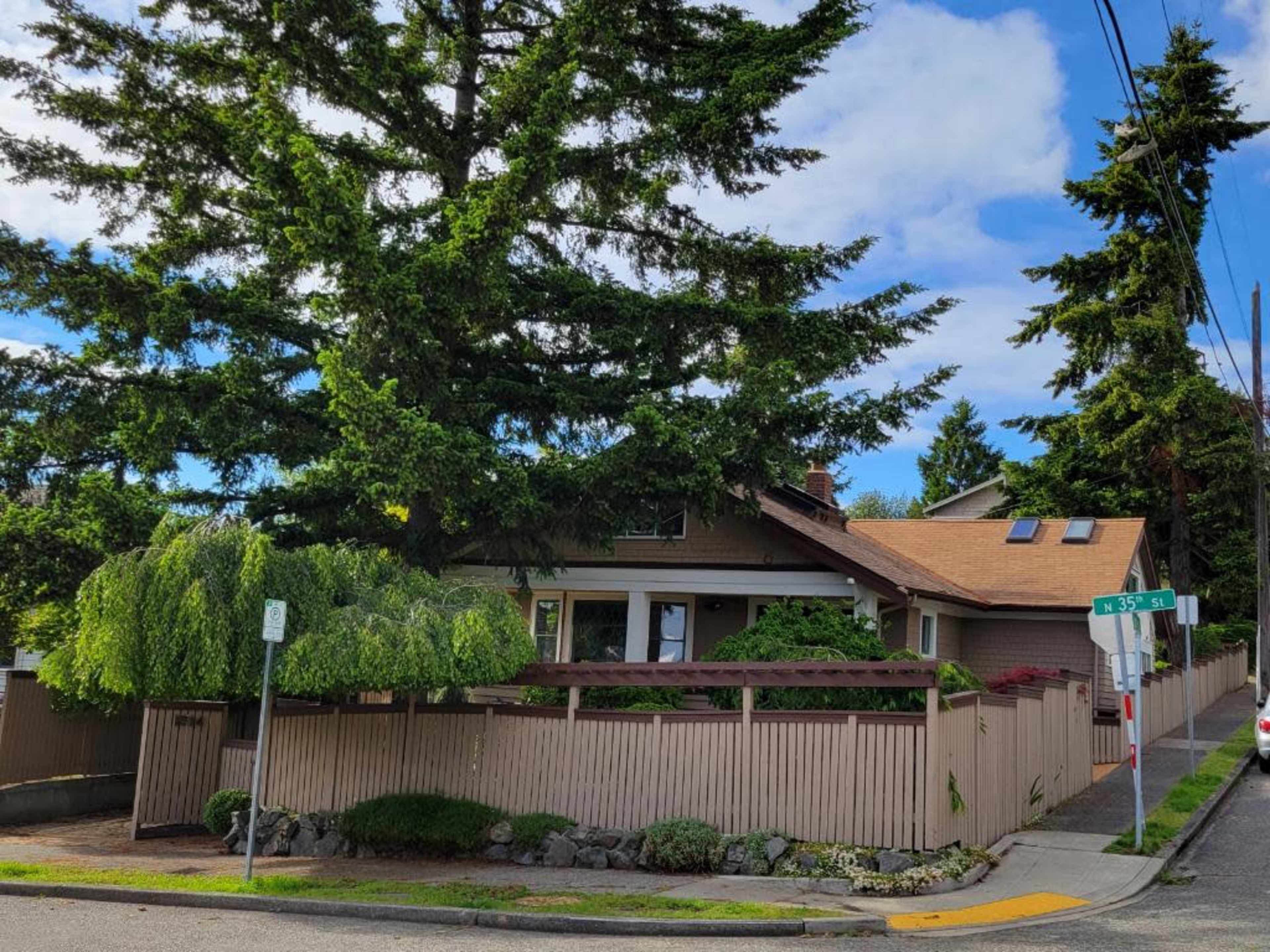 A single-story house with a sloped roof is situated at the corner of a street, surrounded by a wooden fence and tall trees.