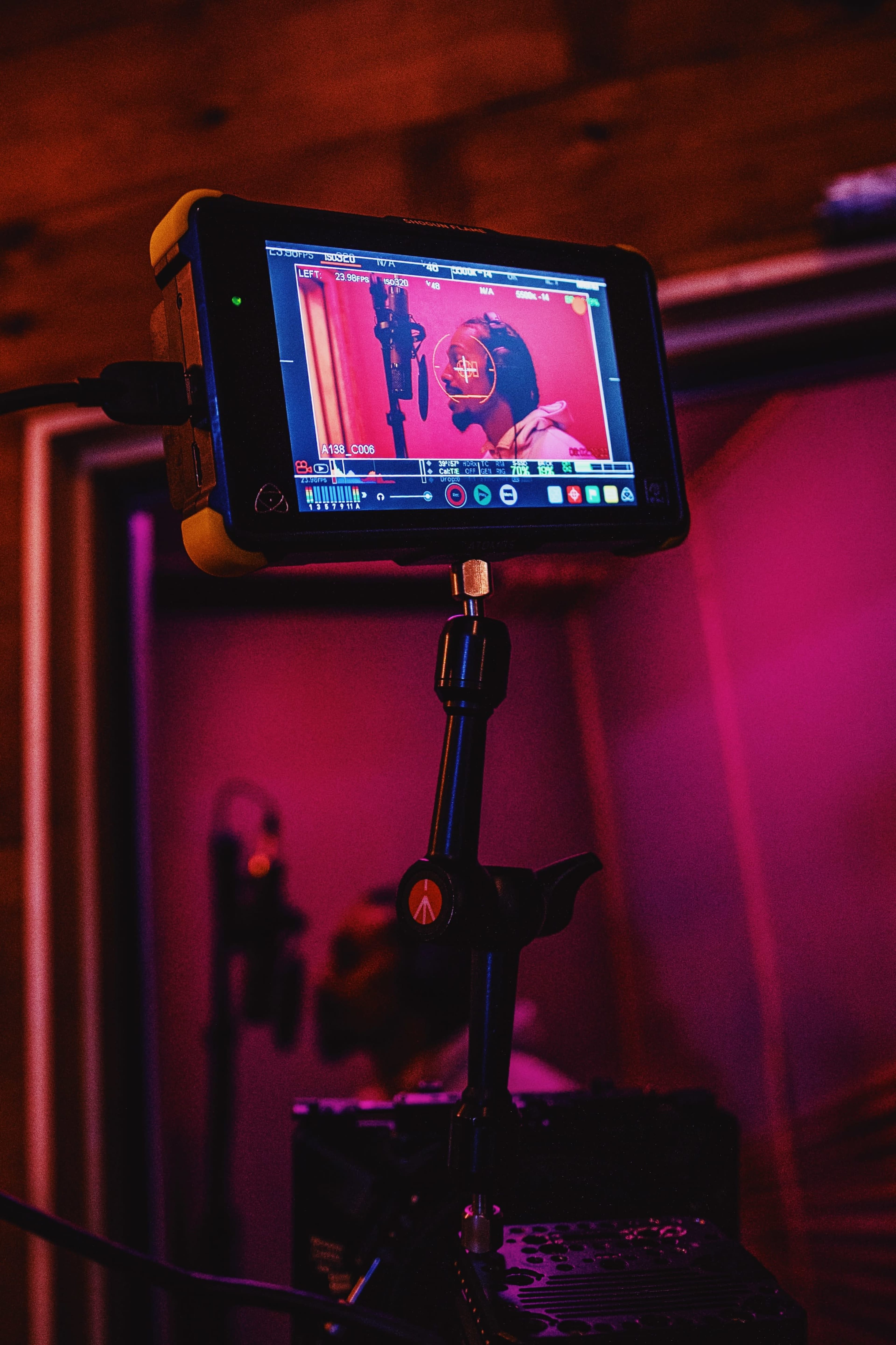 A monitor displaying a recording session in a sound studio is positioned on a stand, showing a microphone and a performer in front of a pink-lit background.