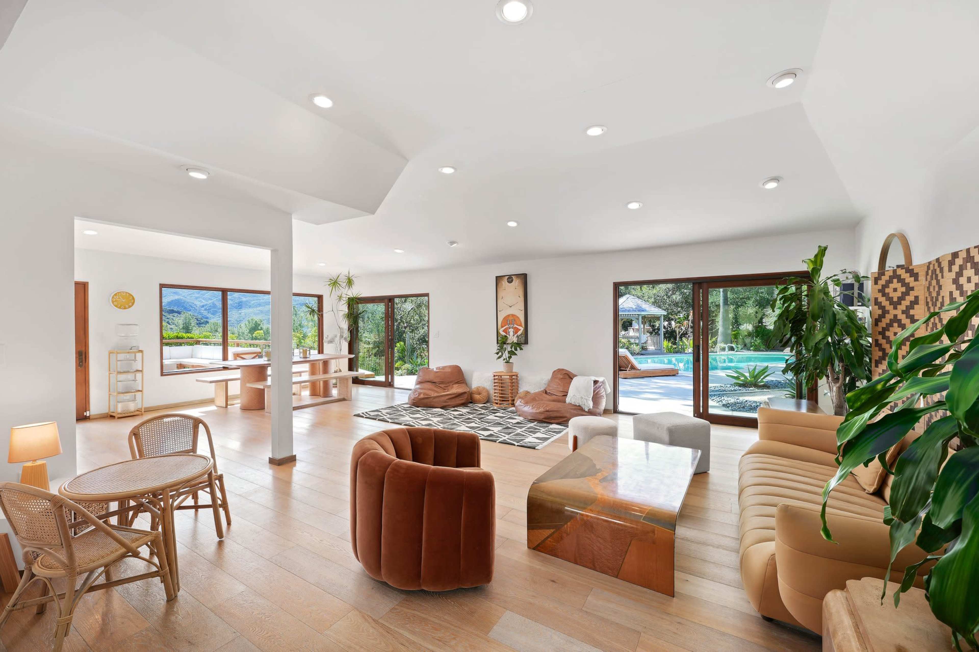 The image features a spacious living room with large windows overlooking a pool and outdoor area, furnished with modern seating and a dining set.