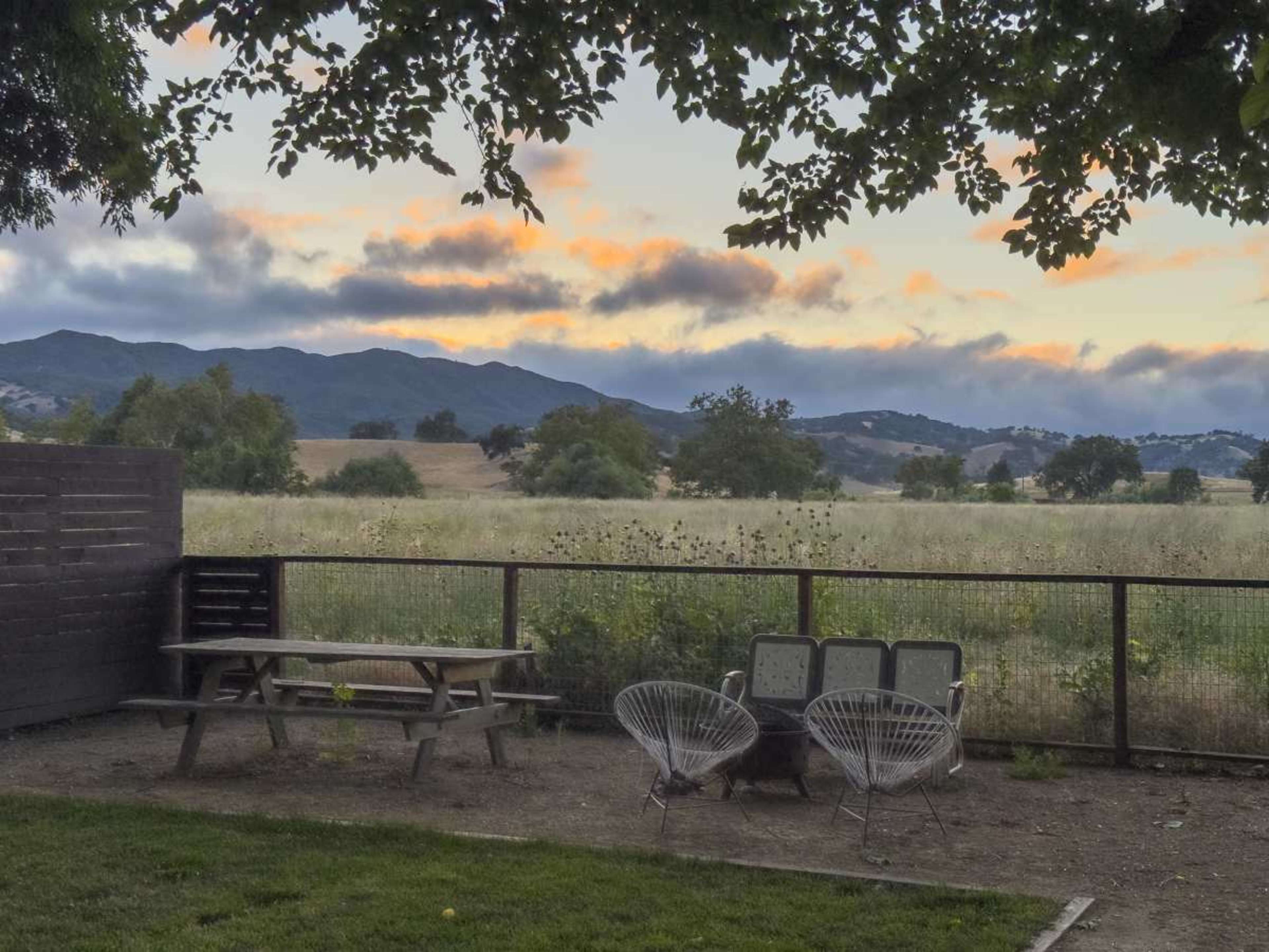 The image shows a backyard scene with a picnic table and two chairs facing a grassy field and rolling hills under a cloudy sky at sunset.