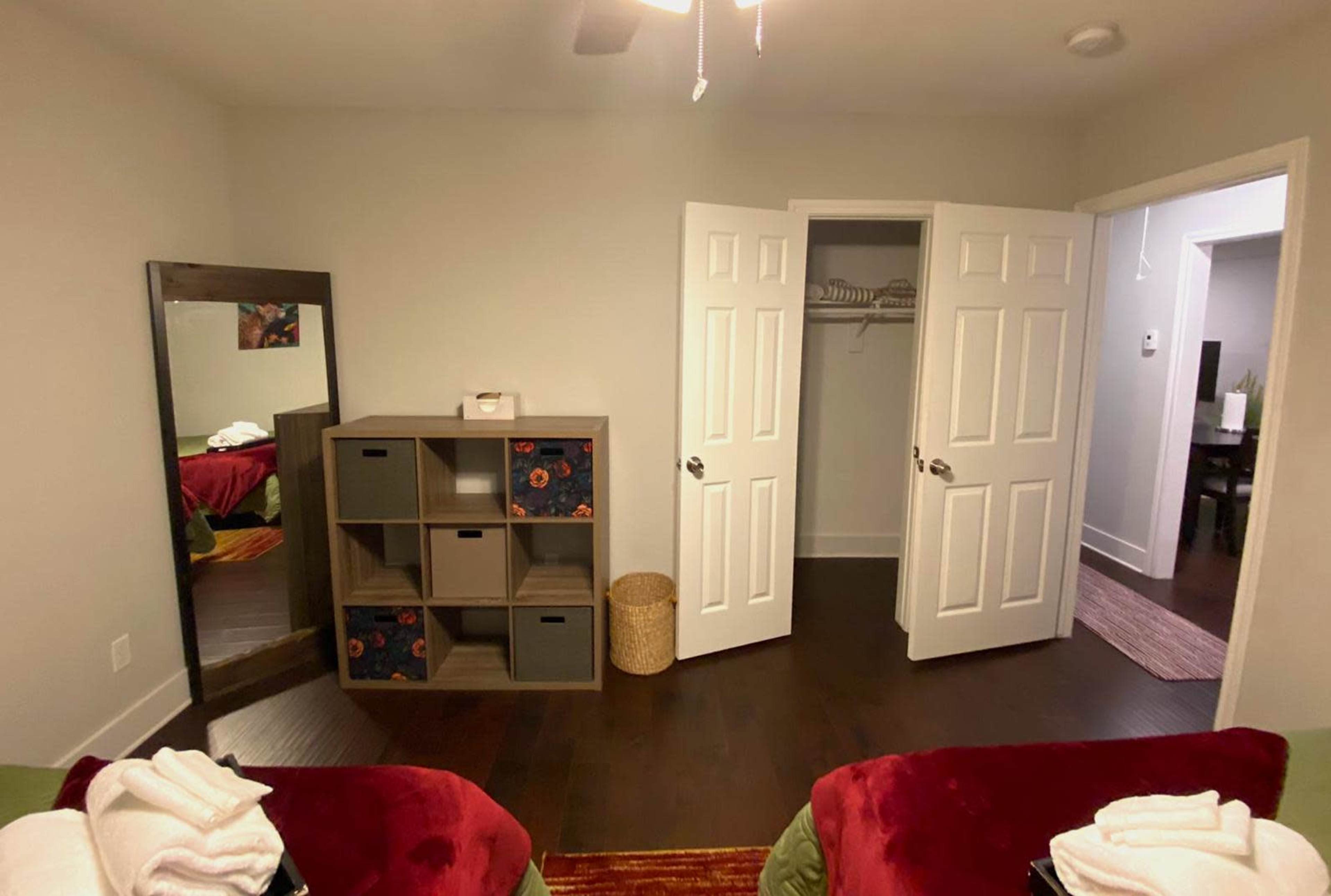 A room features two red chairs, a storage unit with decorative boxes, a large mirror, and an open closet with sliding doors.