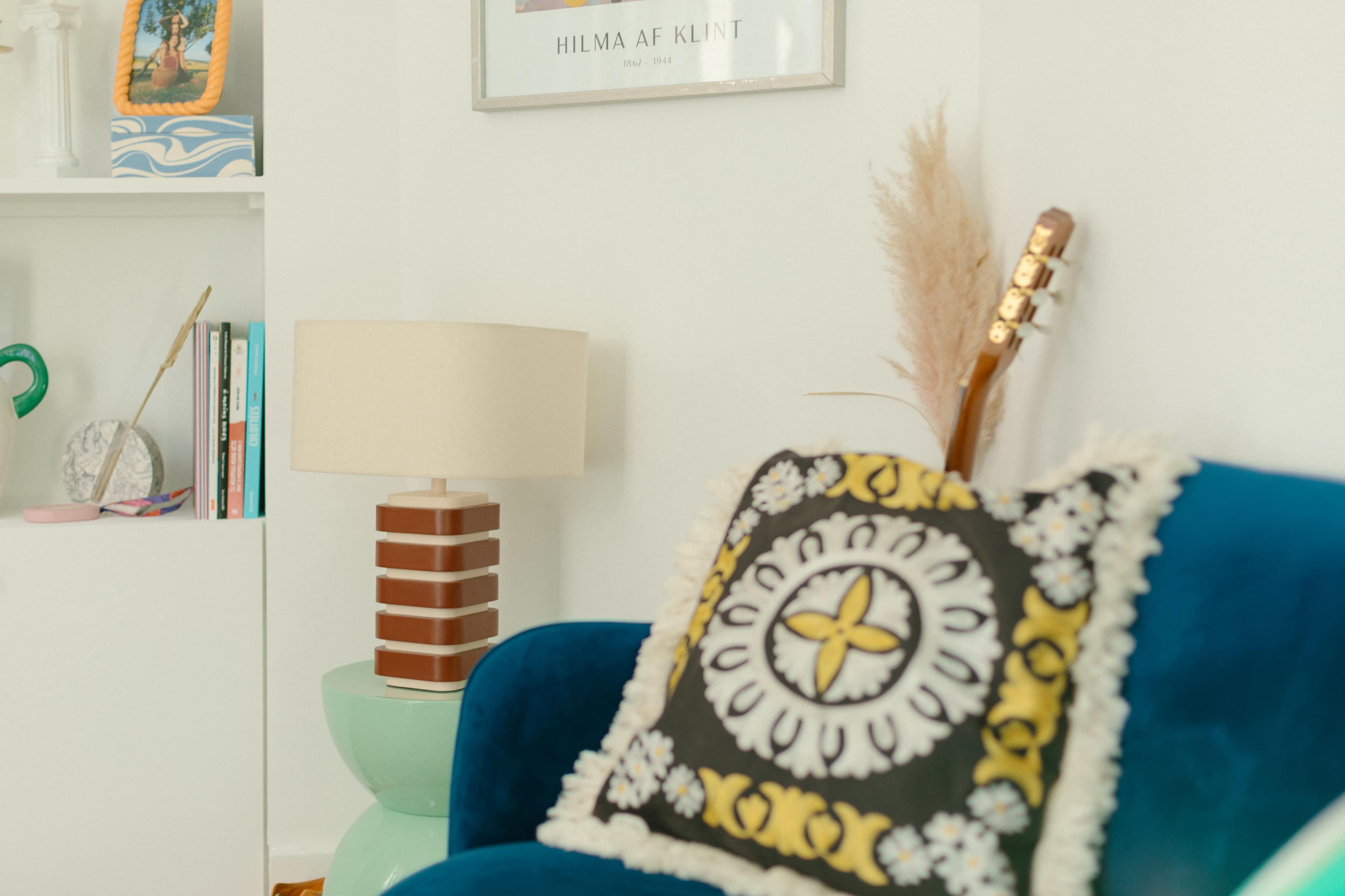 A vibrant living room corner featuring a blue couch with a decorative pillow, a lamp, a guitar resting against the wall, and a shelf with books and decorative items.