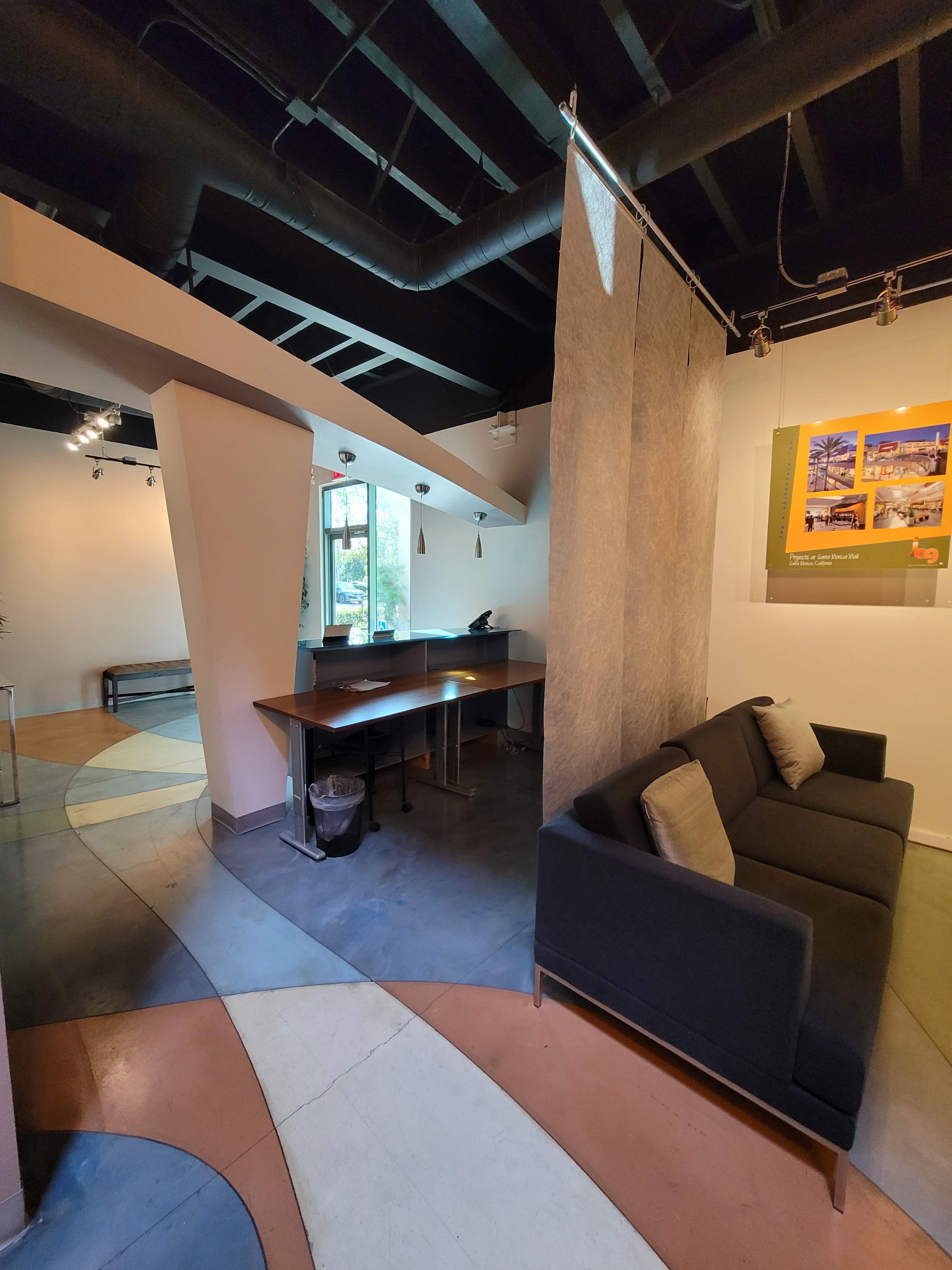A modern office space featuring a black sofa, a desk with a chair, and a partition made of fabric, with unique flooring patterns in muted colors.