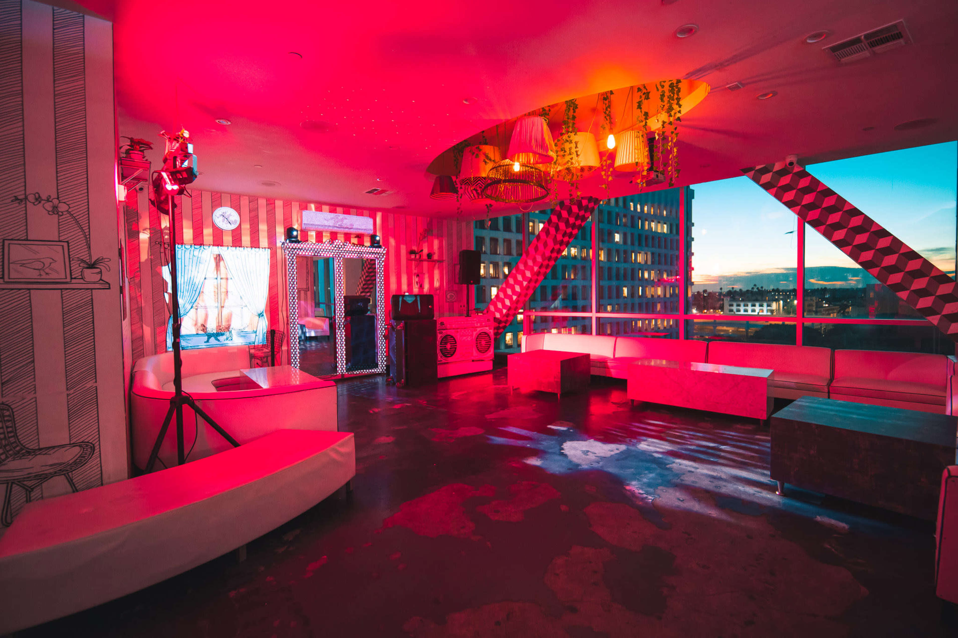 The image shows a brightly lit room with pink and red lighting, featuring modern furniture, large windows, and a concrete floor.