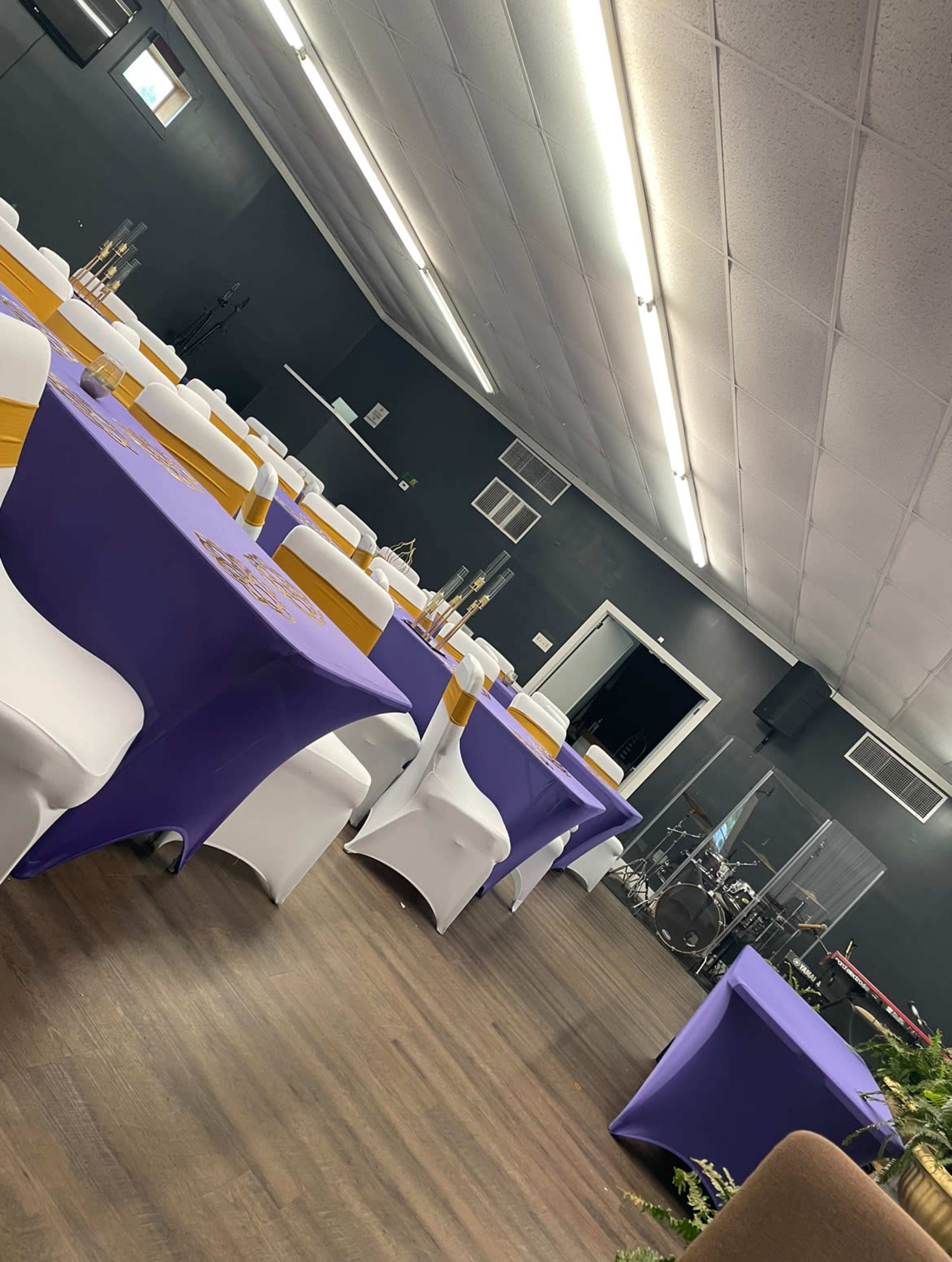 A room set up for an event with purple and white tablecloths, wooden flooring, and chairs arranged around the tables, with a stage and musical instruments visible in the background.