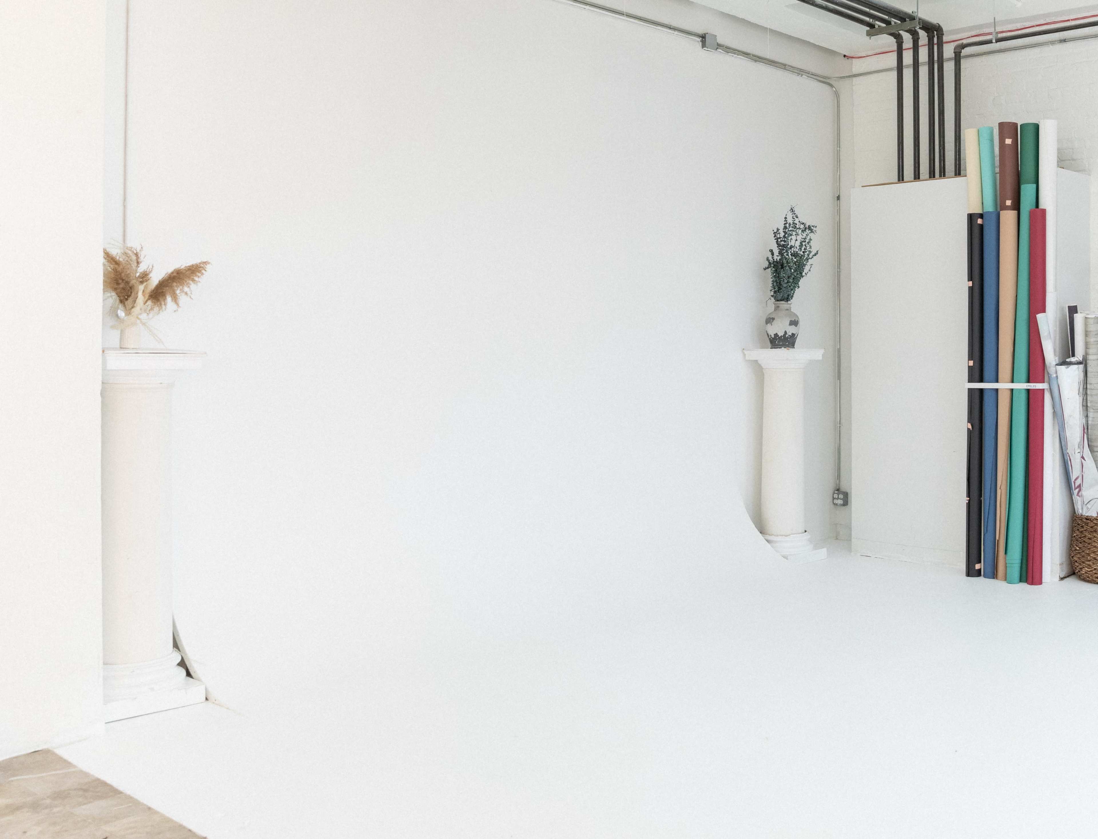 A minimalist studio space features white walls and flooring, with two decorative columns holding plants and various rolls of colored paper leaning against the wall.
