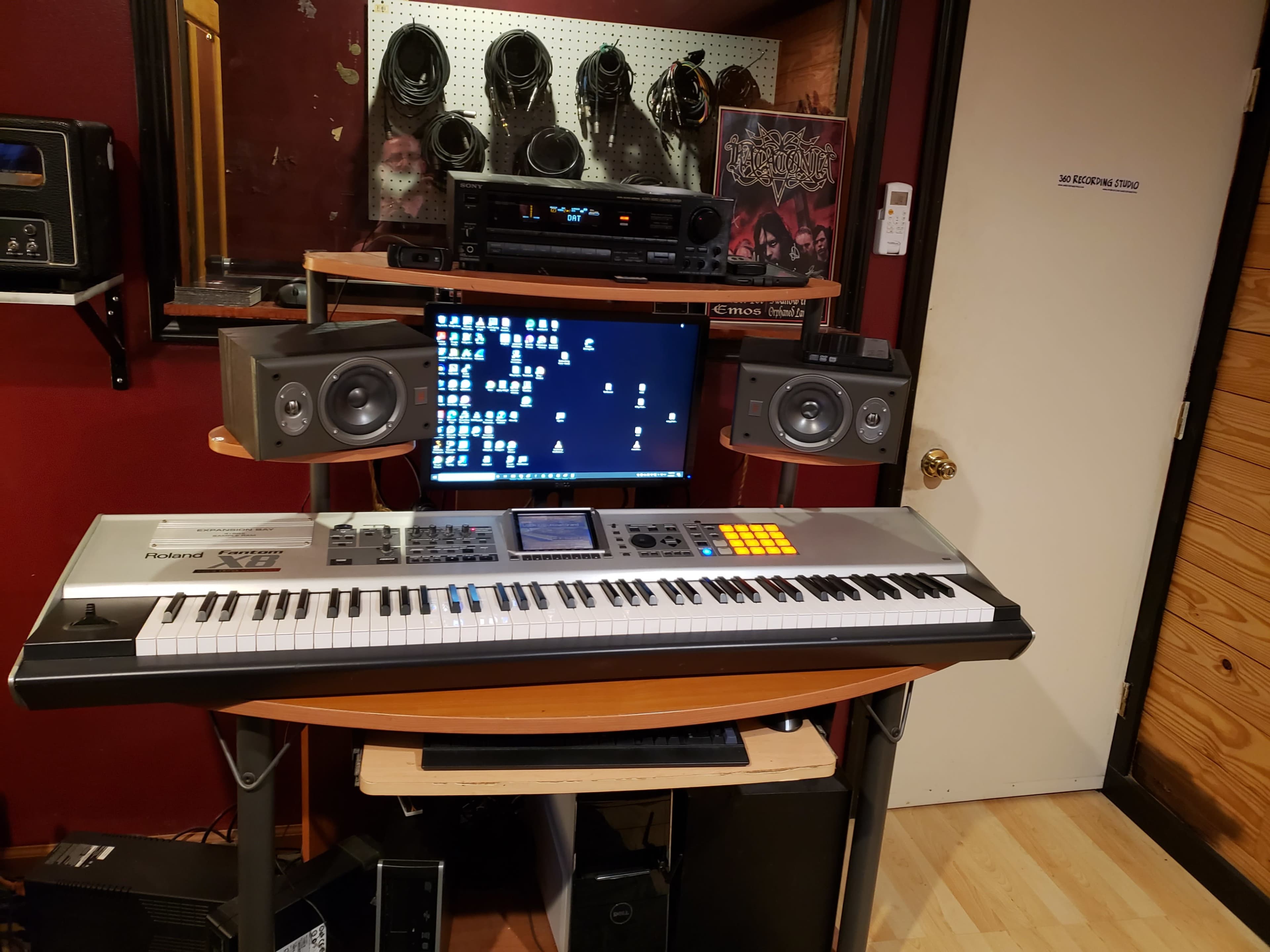 A music production setup featuring a keyboard, computer monitor, and audio speakers arranged on a desk against a red wall.