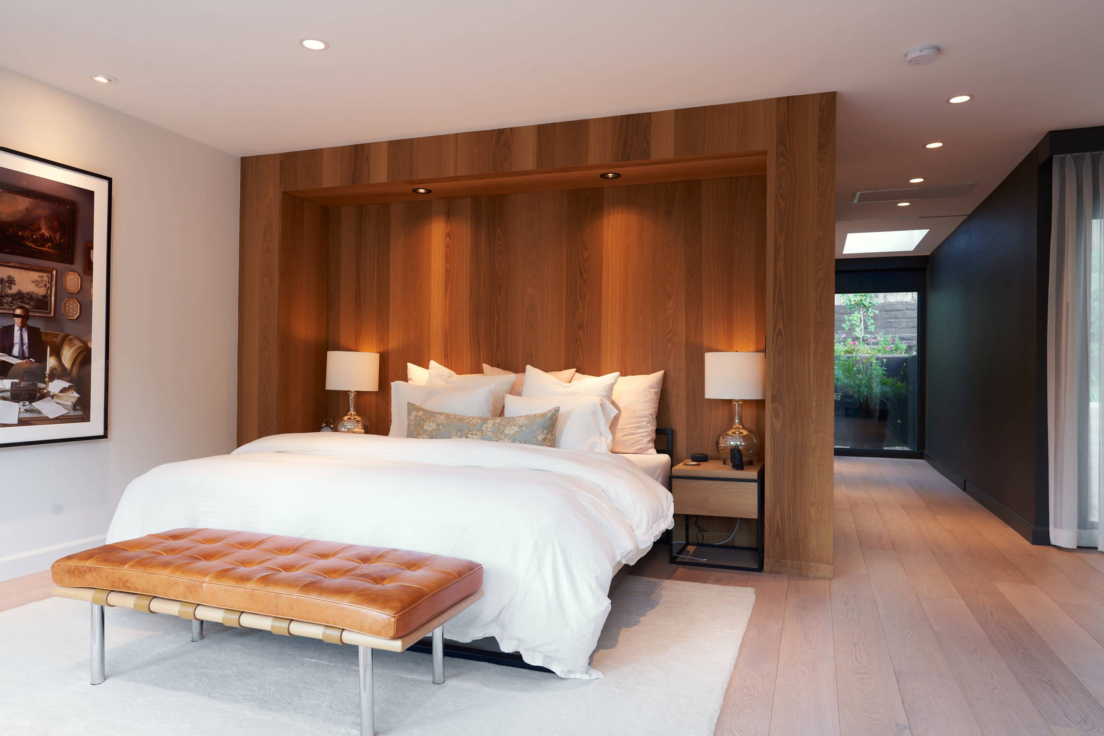 A modern bedroom with a wooden accent wall, a white bed with multiple pillows, a brown leather bench at the foot of the bed, and two bedside lamps.