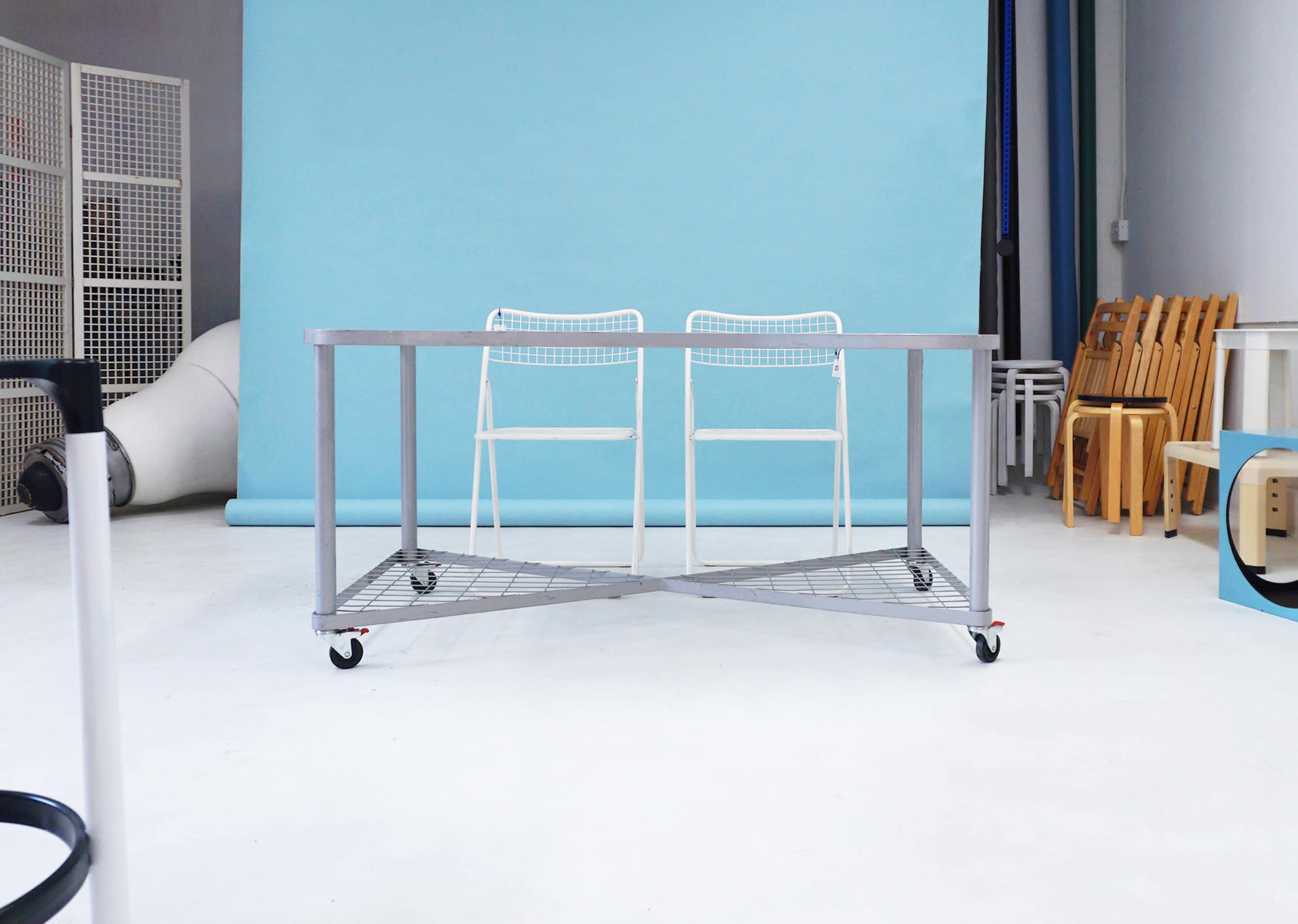 A metal table with a wire mesh bottom and two white chairs is positioned in front of a light blue backdrop in a studio setting.