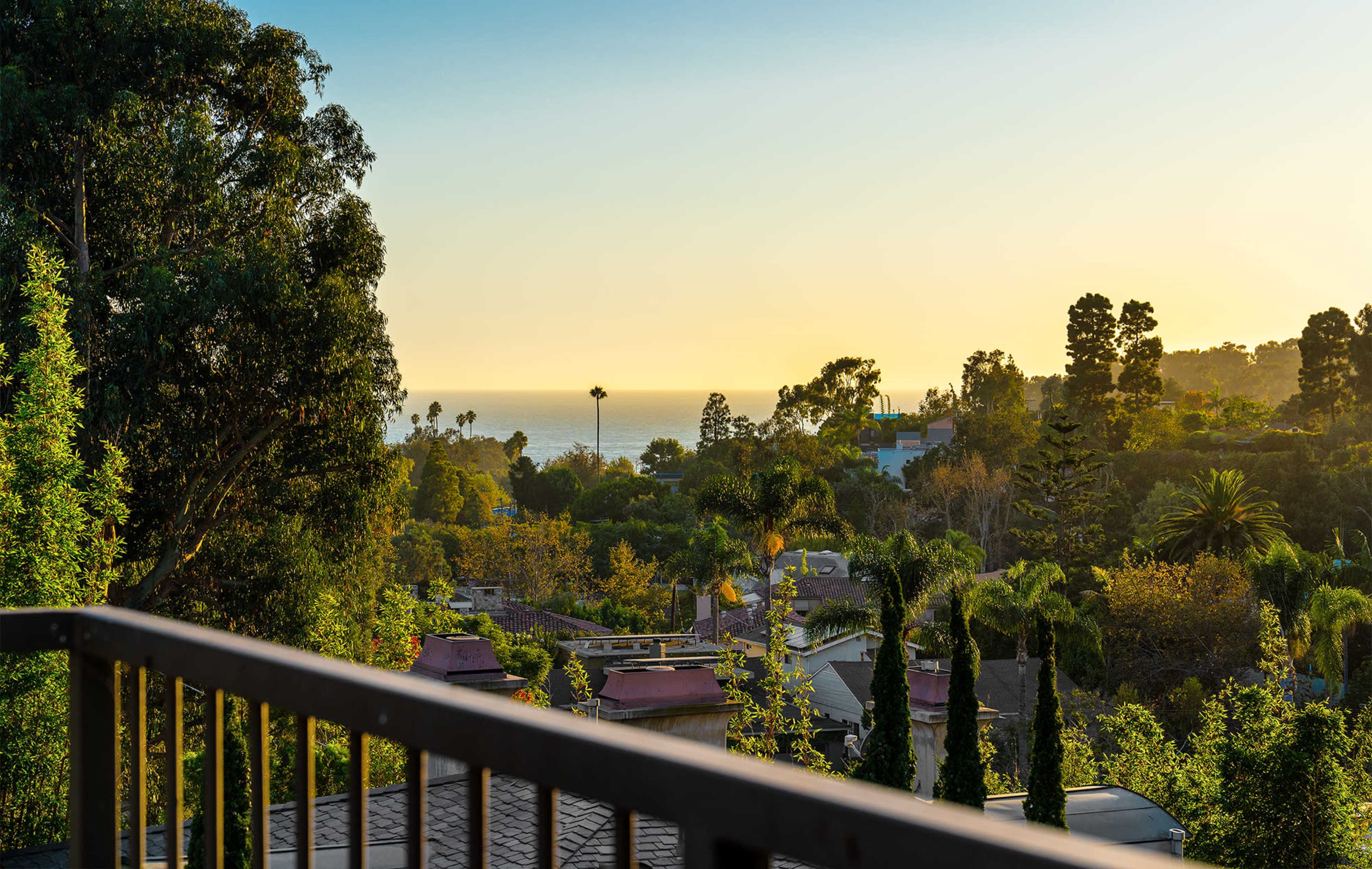 A coastal view at sunset, framed by trees and residential rooftops.