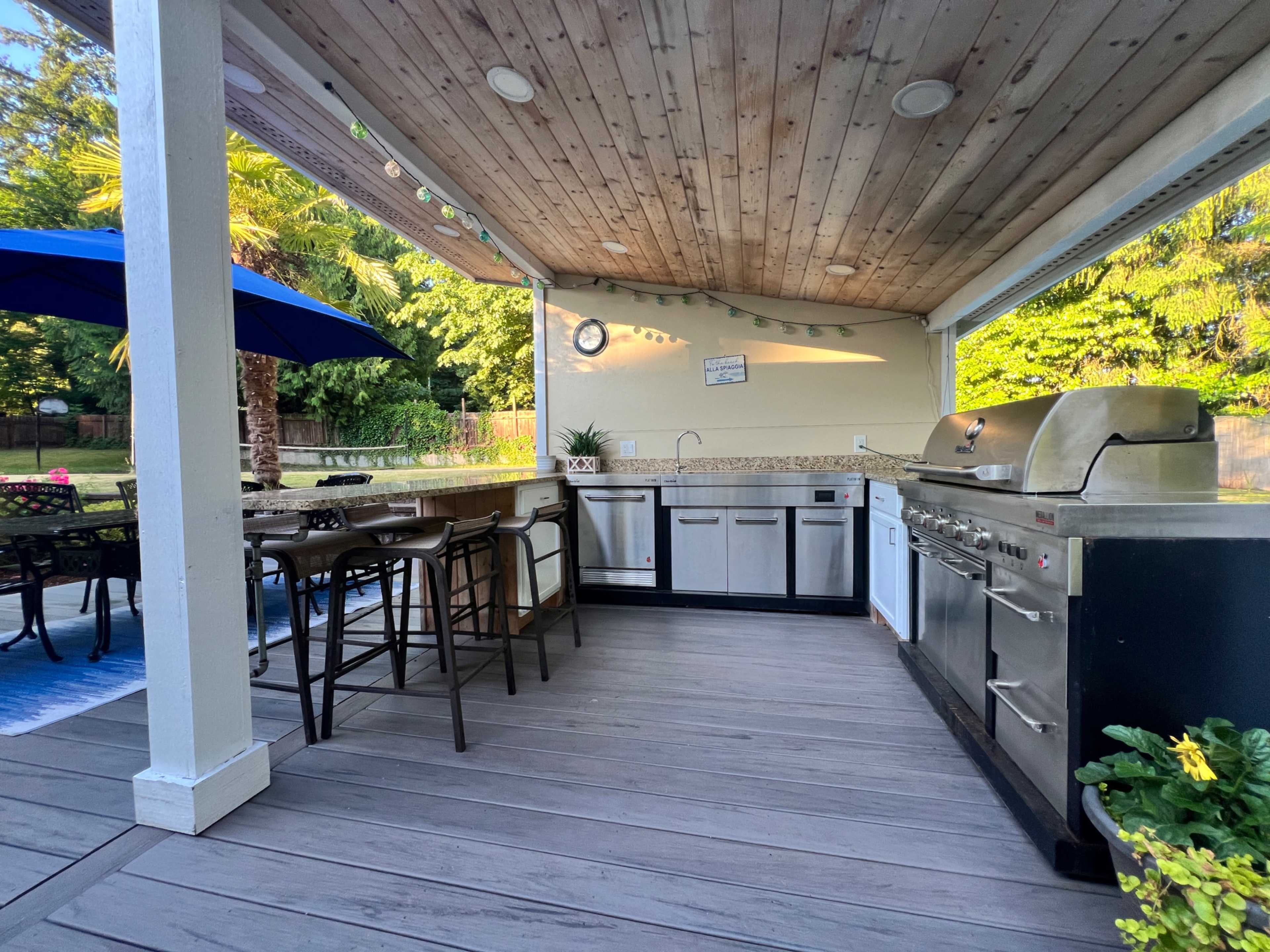 The image shows an outdoor kitchen featuring a grill, countertop, bar stools, and a shaded seating area with a blue umbrella.