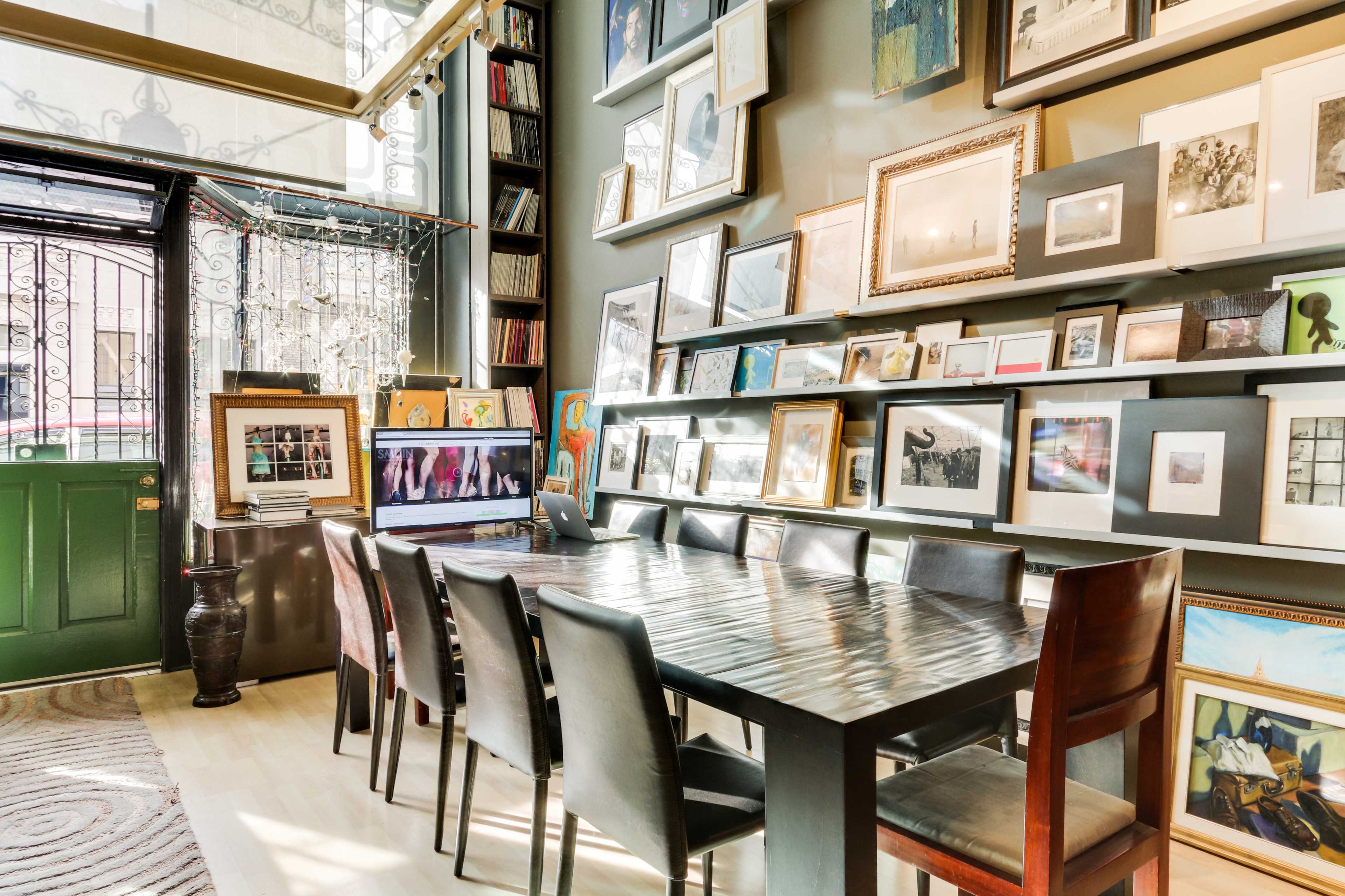 A long wooden table with several black chairs is set in a room filled with framed photographs and artwork on the walls, with large windows letting in natural light.