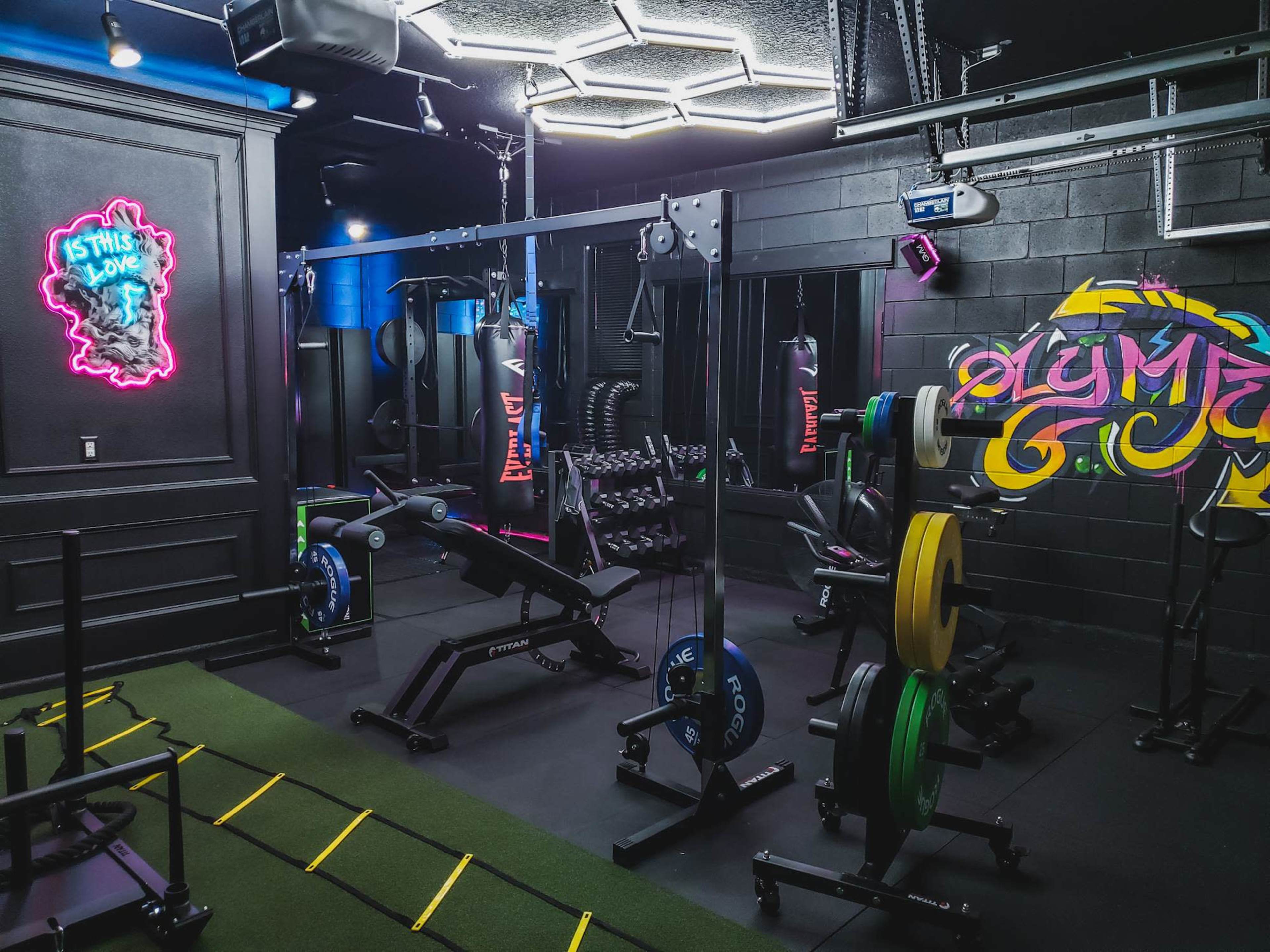 The image shows a modern gym interior equipped with various weightlifting machines, free weights, and a colorful neon sign on the wall.