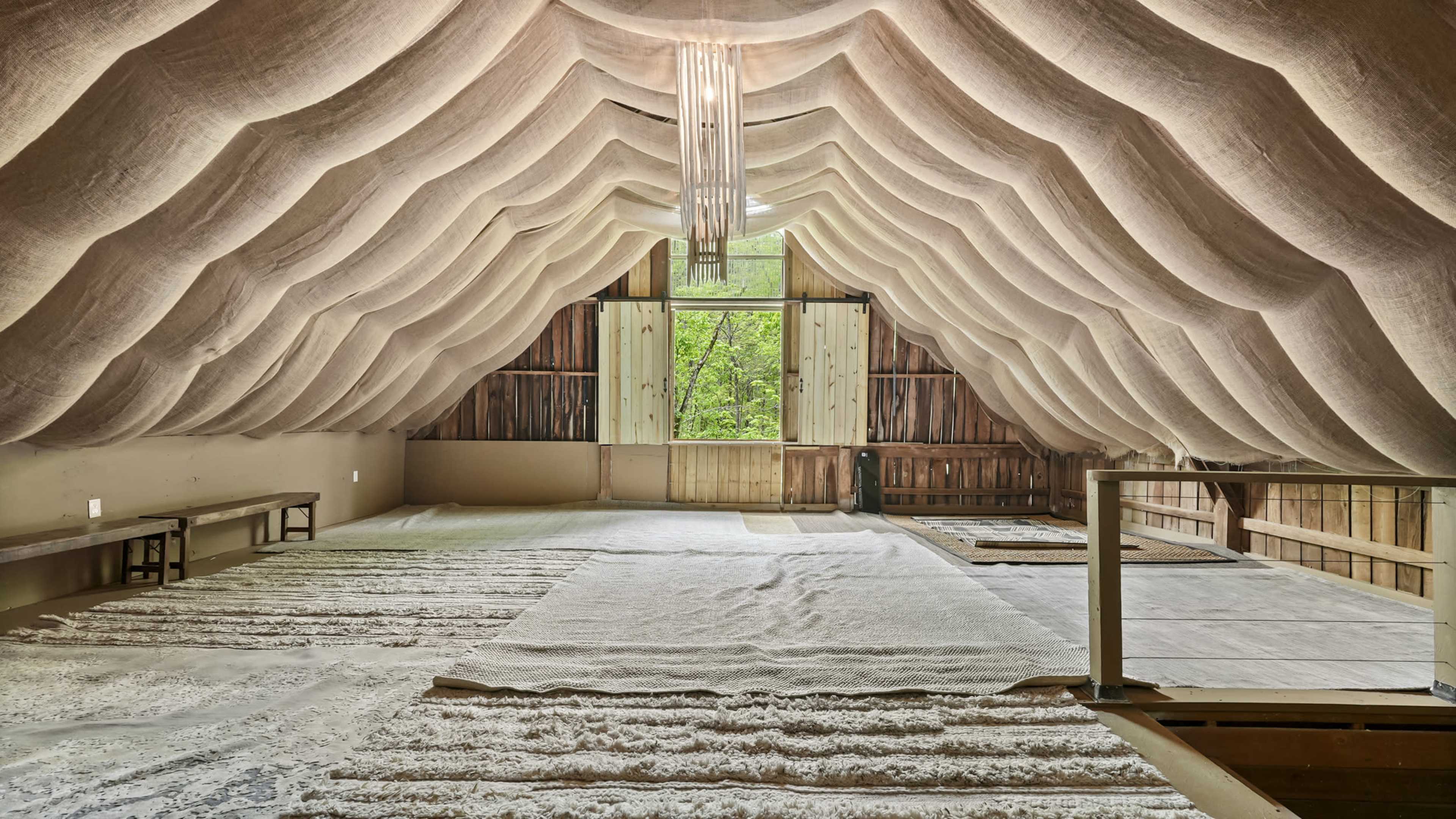 The image shows an empty room with a high, textured ceiling and large windows, featuring natural light and wooden elements.