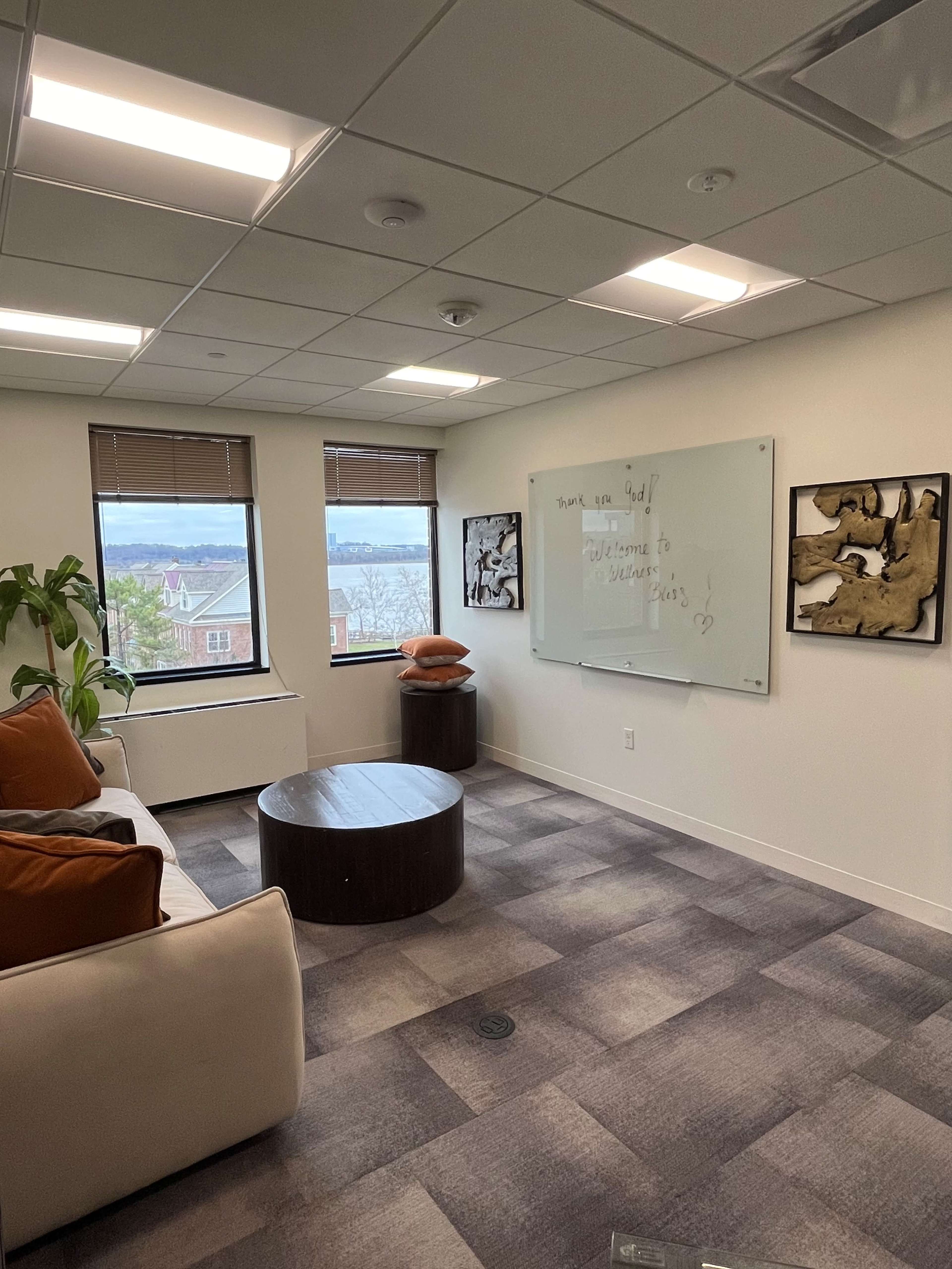 A modern office with a seating area, a round coffee table, a whiteboard, and large windows overlooking a body of water.