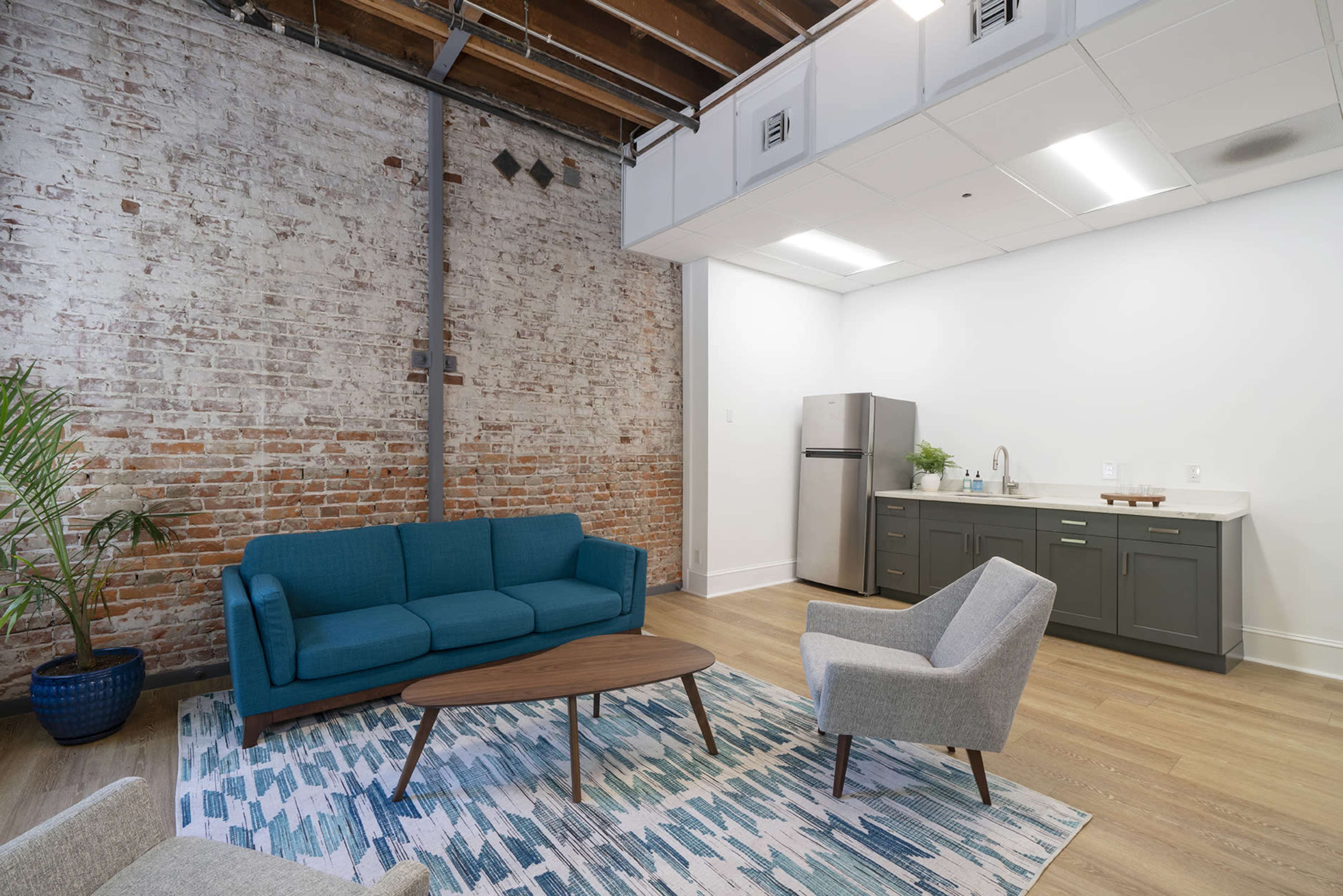 A modern room featuring a blue sofa, a gray chair, and a wooden coffee table on a patterned rug, with exposed brick walls and a kitchenette in the background.