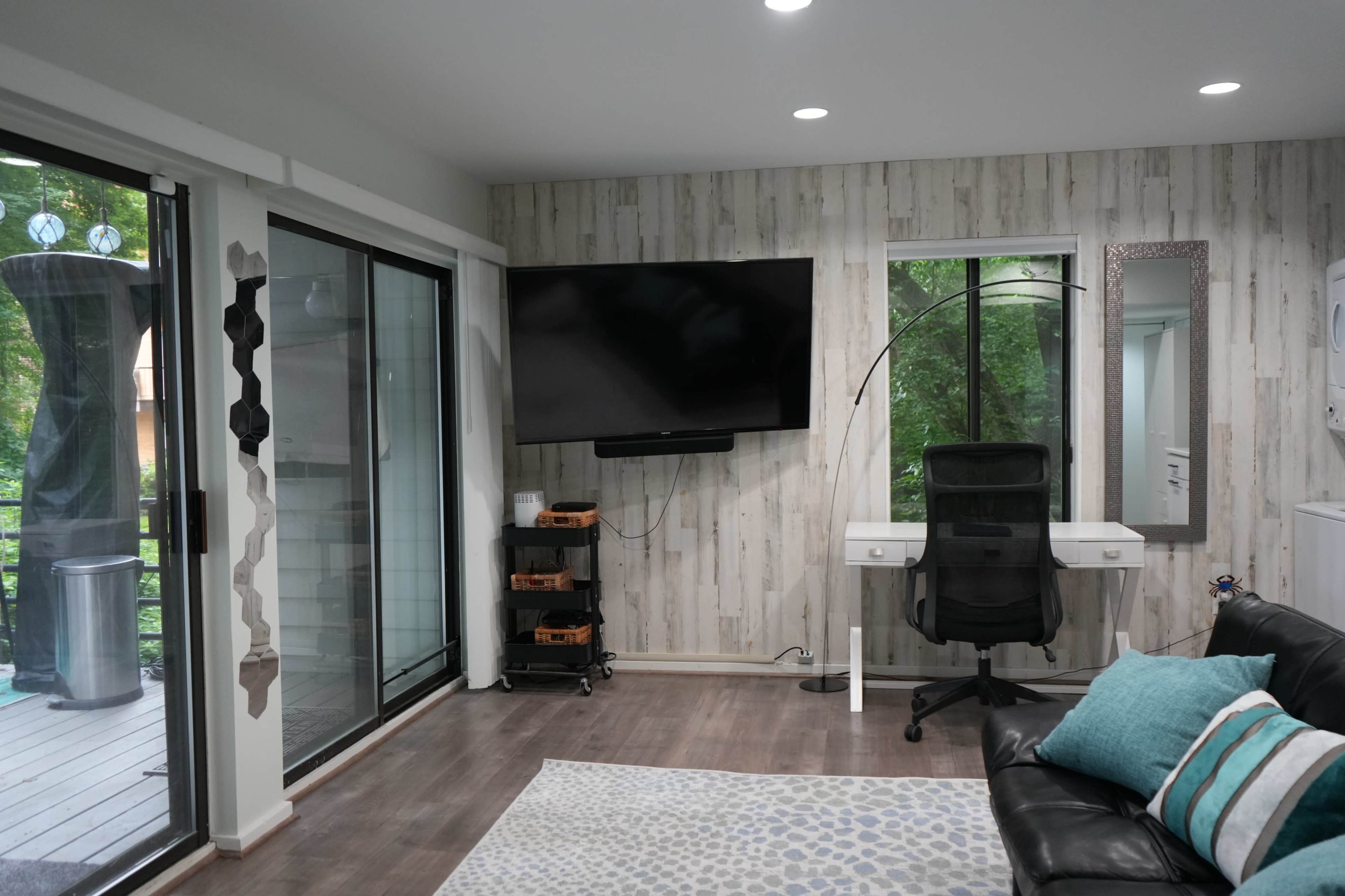 The image shows a modern living space with a black leather couch, a large wall-mounted TV, a desk with a chair, and sliding glass doors that lead to an outdoor area.