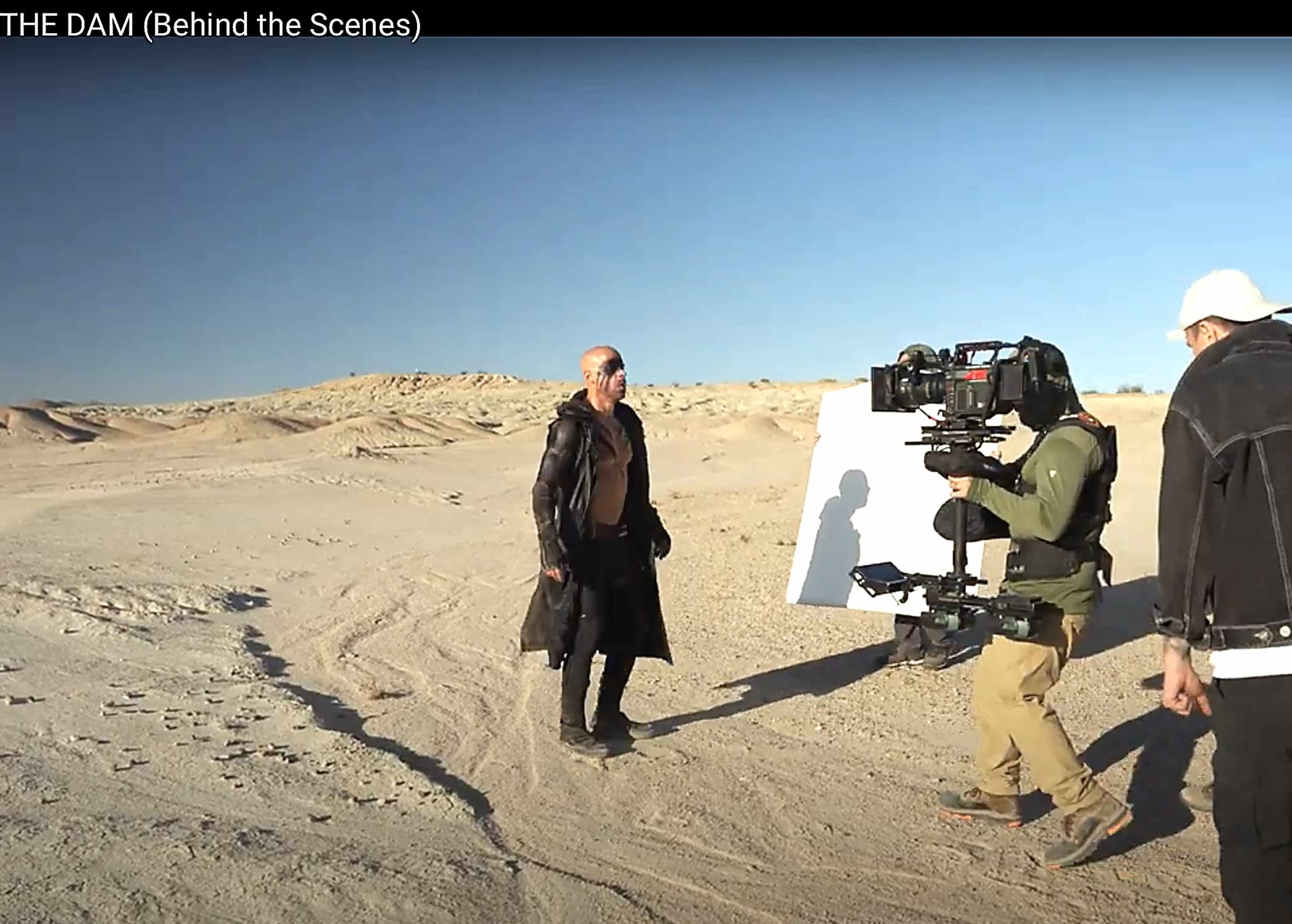 A man in a black coat stands in a desert landscape while a crew member operates a camera on a stabilizing rig.