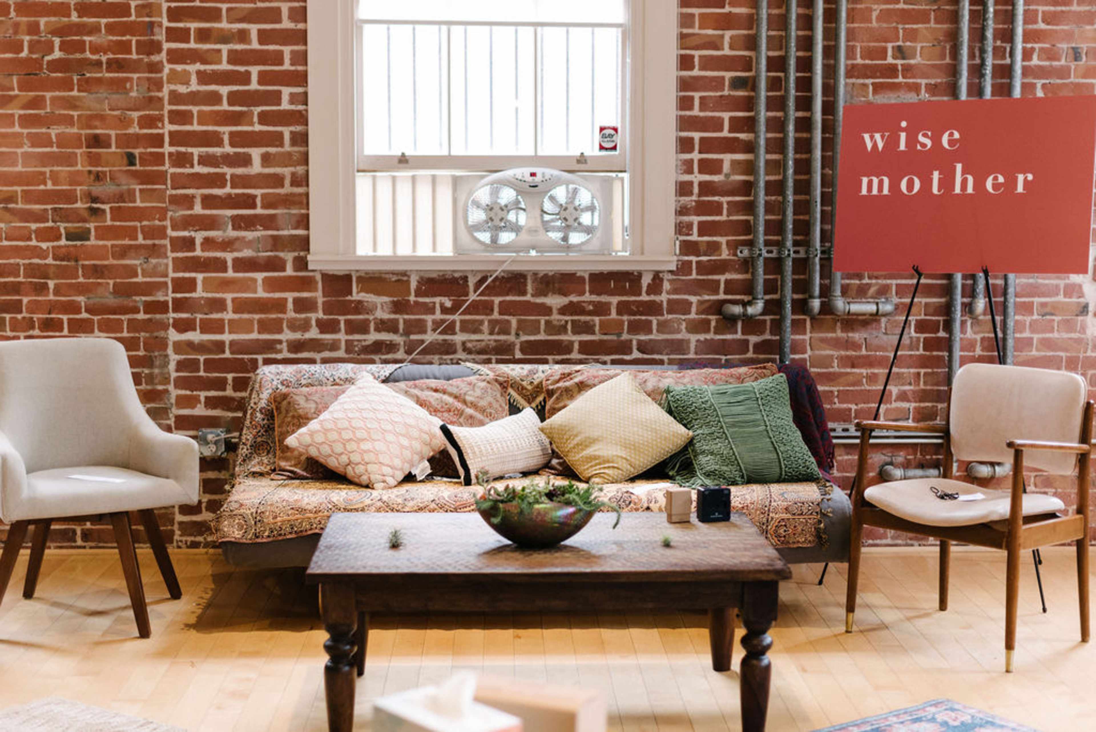 The scene features a cozy sitting area with a patterned sofa adorned with multiple pillows, a wooden coffee table, and a sign reading "wise mother" against a brick wall.