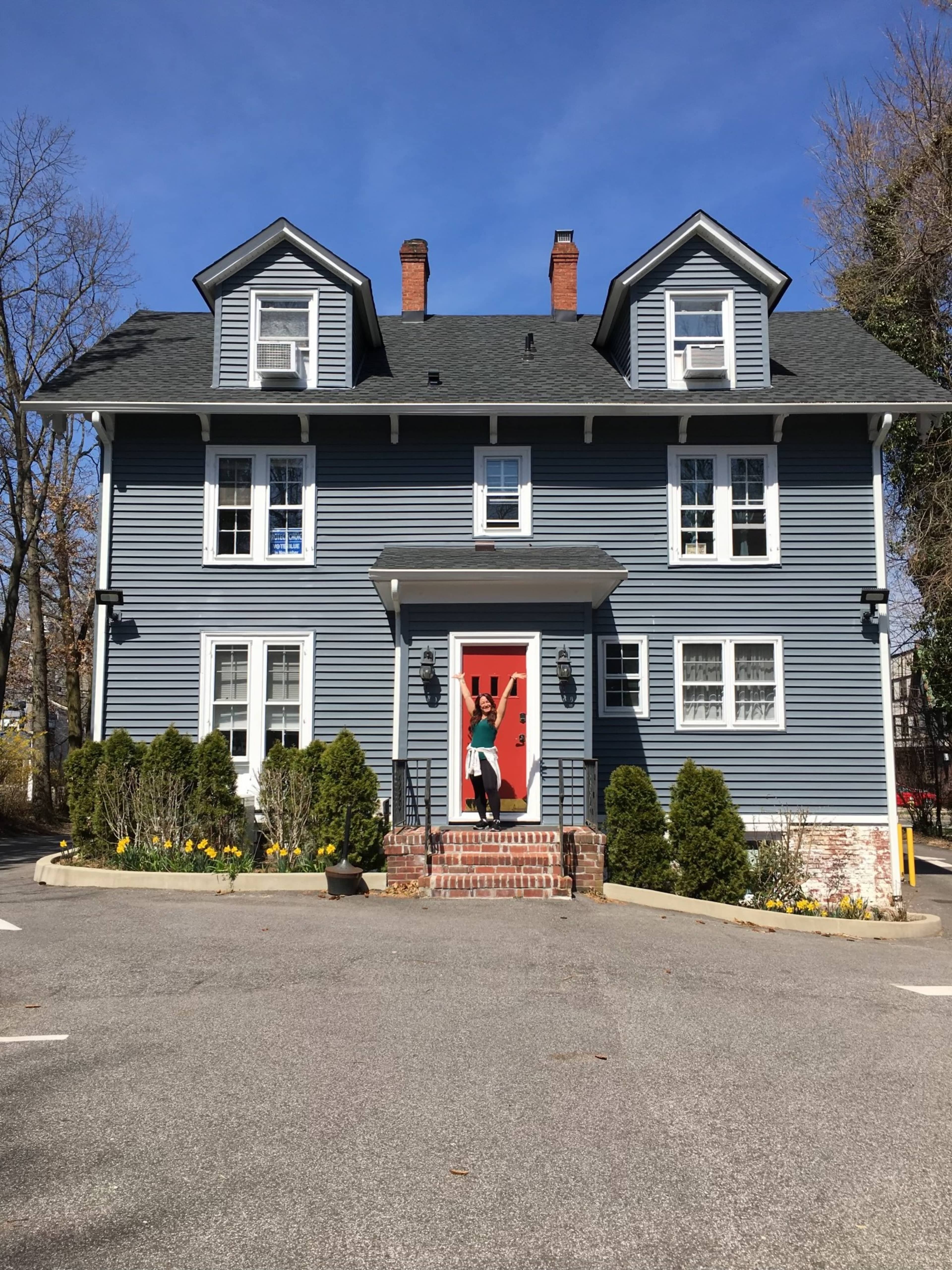 A blue two-story house with a red front door and symmetrical windows sits on a paved area surrounded by green shrubs and flowers.