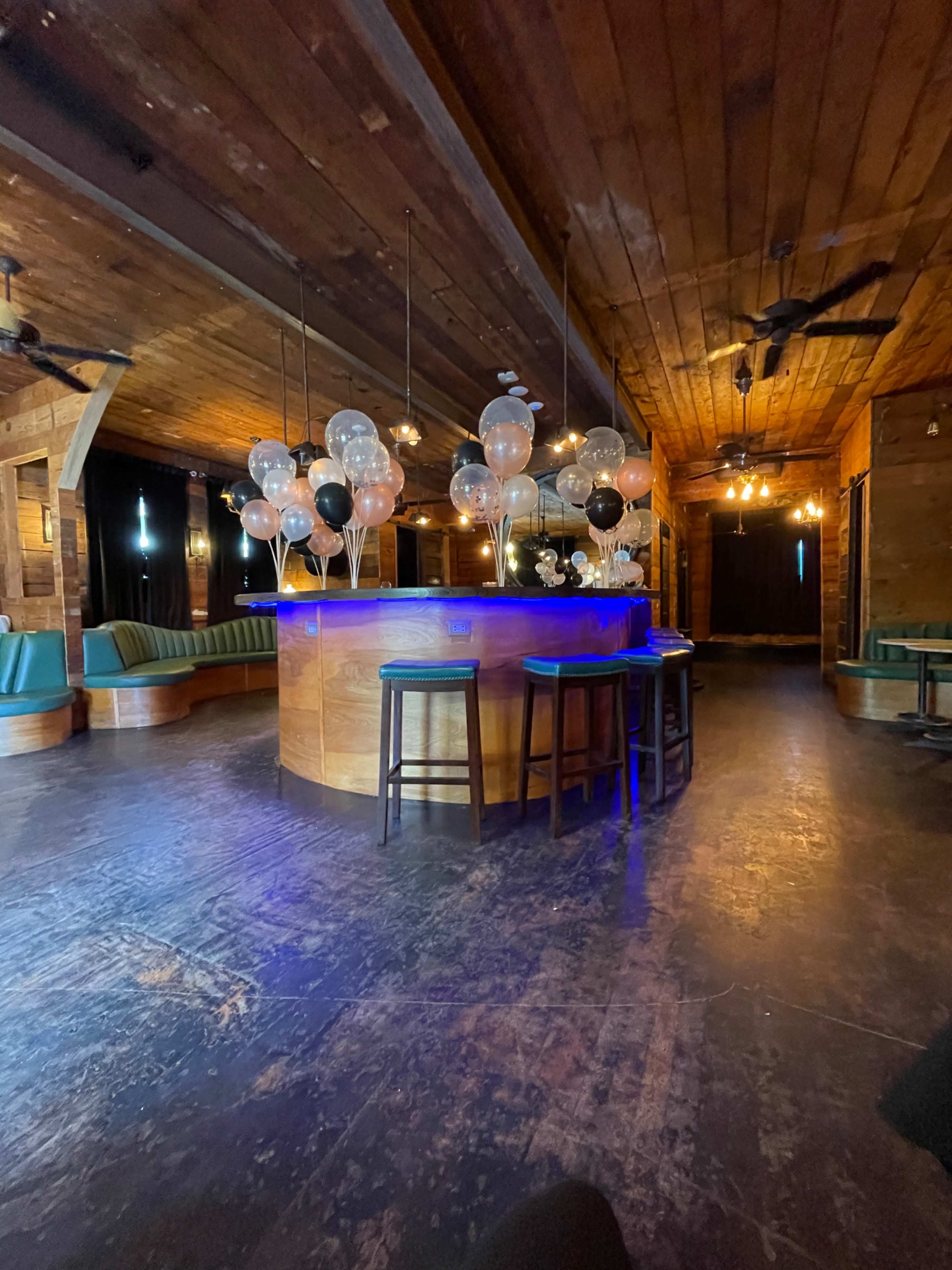 A bar area with a rounded wooden counter, surrounded by black and white balloons, in a dimly lit space with wooden walls and a seating area.
