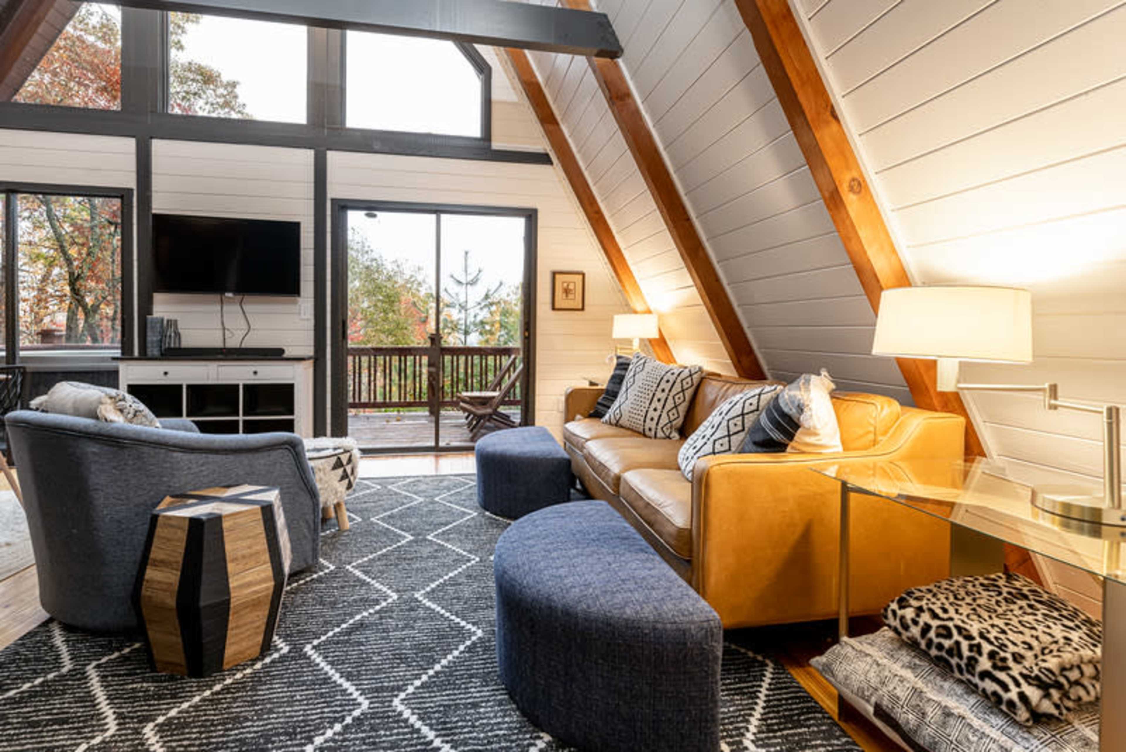 The interior of a modern A-frame living room features a leather couch, armchairs, a coffee table, and large windows opening to a deck with a view of trees.