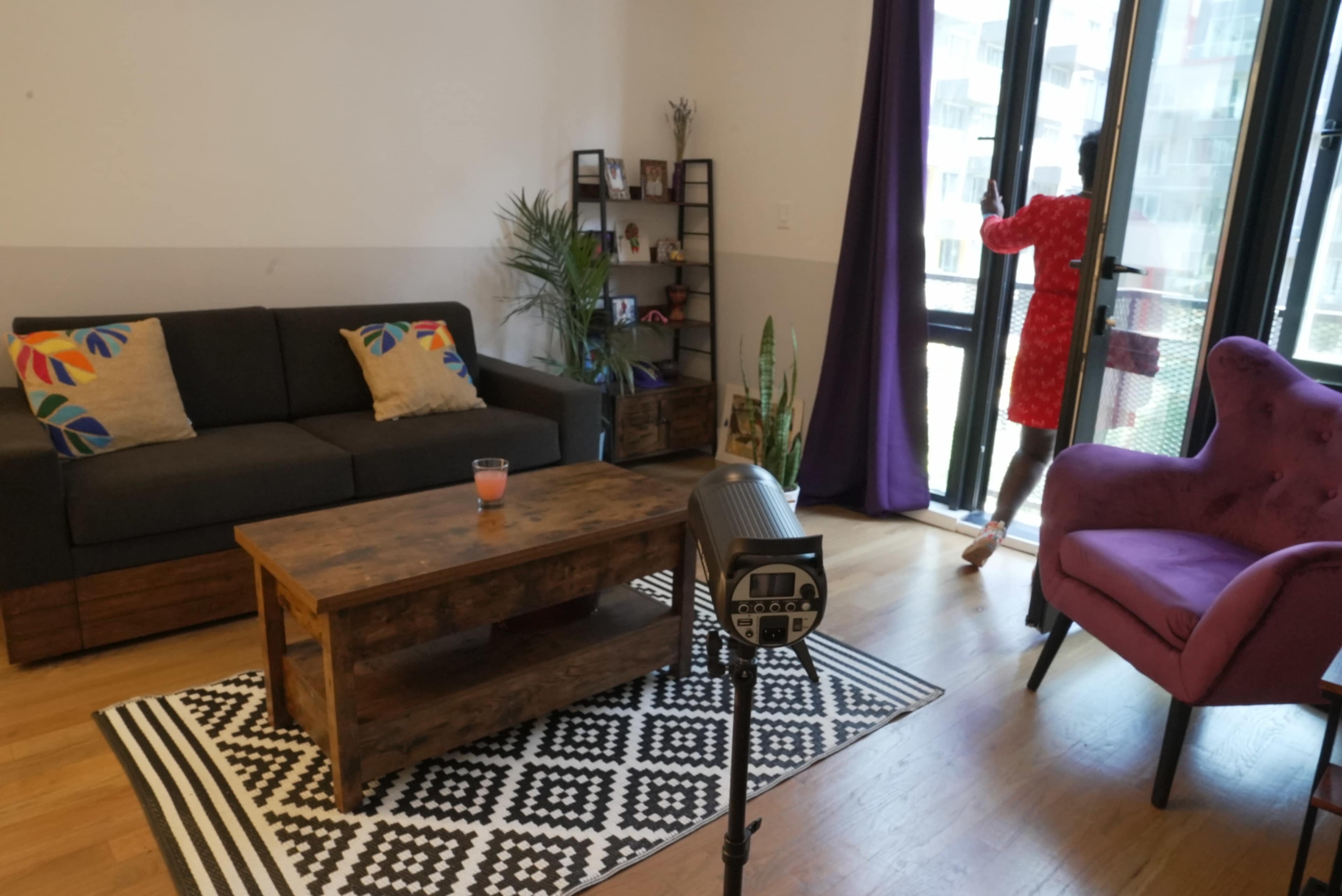 A living room features a dark sofa, a purple chair, a wooden coffee table, and a person in a red dress standing by a sliding glass door.