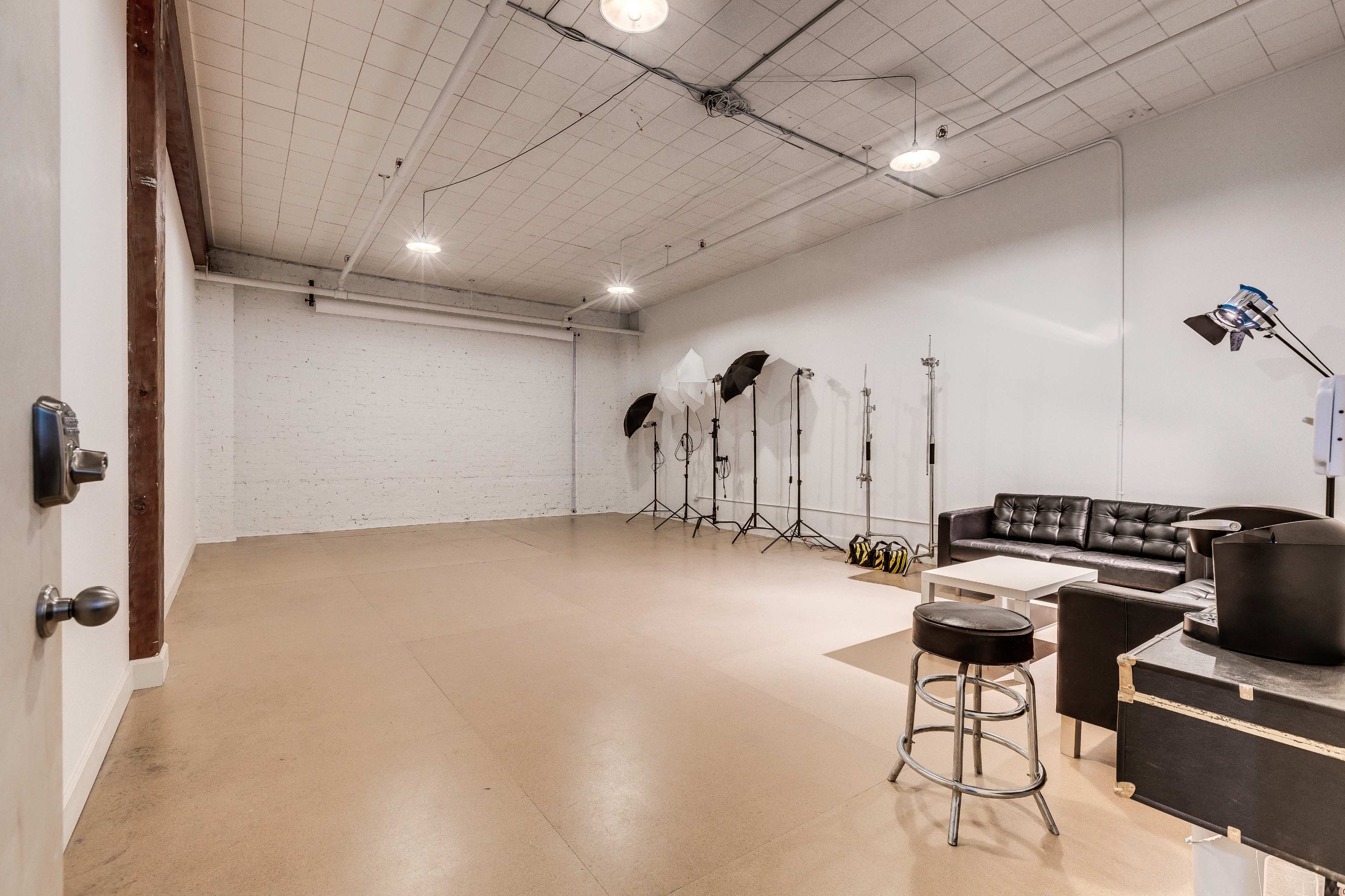 A spacious studio with a bare concrete floor, a black leather couch, and several photography lighting equipment setups against a white wall.